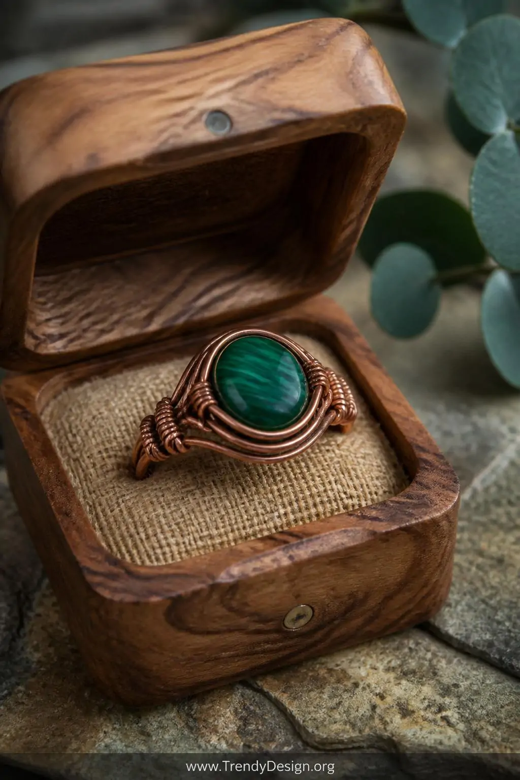 Handmade copper wire ring with a deep green malachite cabochon stone setting resting inside an open raw wood ring box on a stone surface, earthy greens and warm copper tones, soft natural light, extreme closeup shot