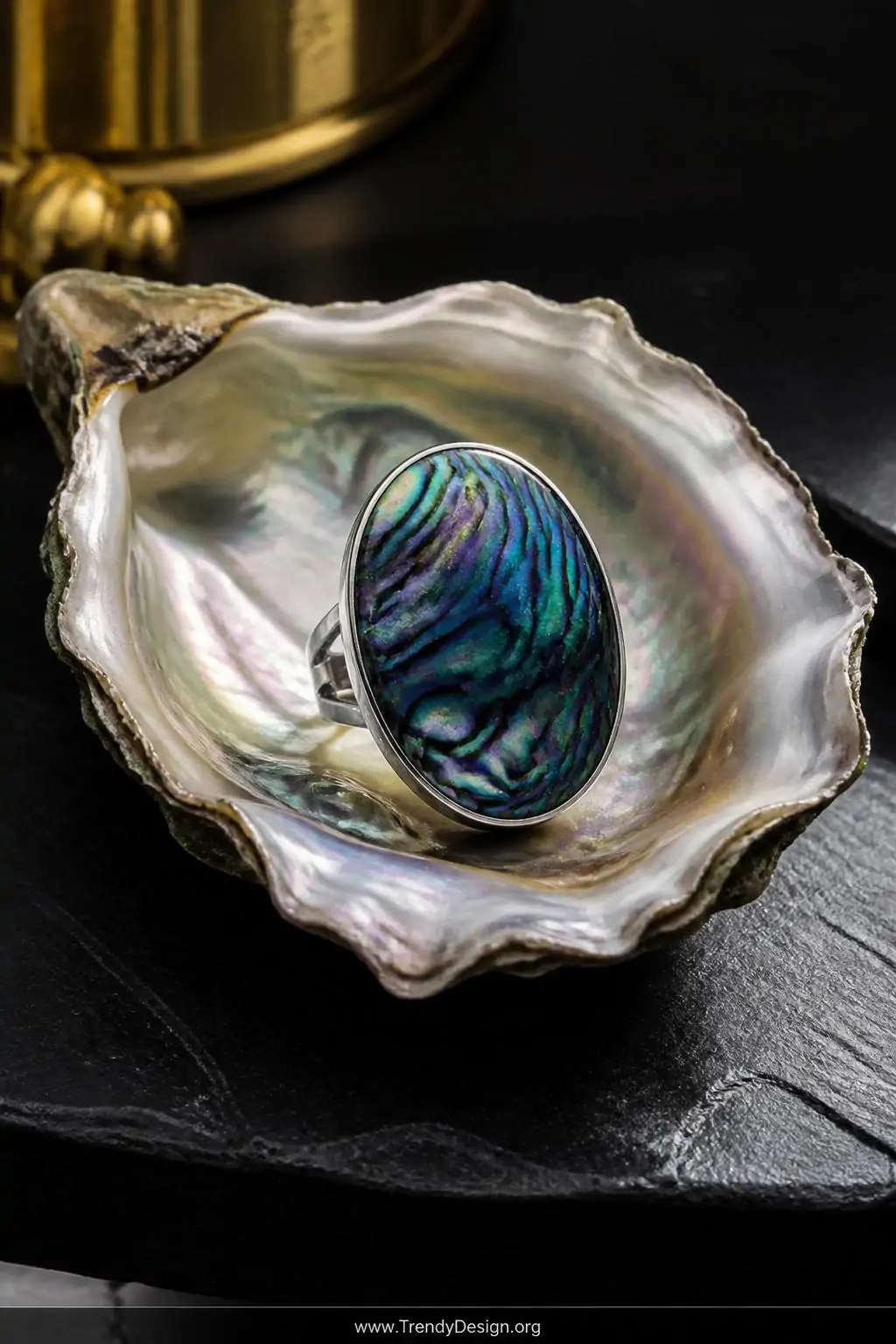 Iridescent abalone shell cocktail ring with glass-smooth resin setting resting inside an open oyster shell on a dark slate vanity surface, dramatic moody studio lighting catching blue green purple swirls, closeup shot, photorealistic interior design