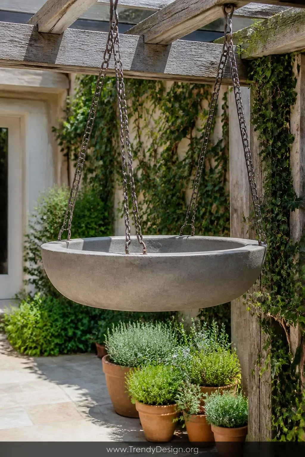 Hanging shallow concrete basin suspended by heavy brushed iron chains from a weathered pergola beam over a compact stone patio, smooth cool gray concrete contrasting against the delicate metal links, surrounded by trailing ivy and small potted herbs below, medium shot in bright midday light.