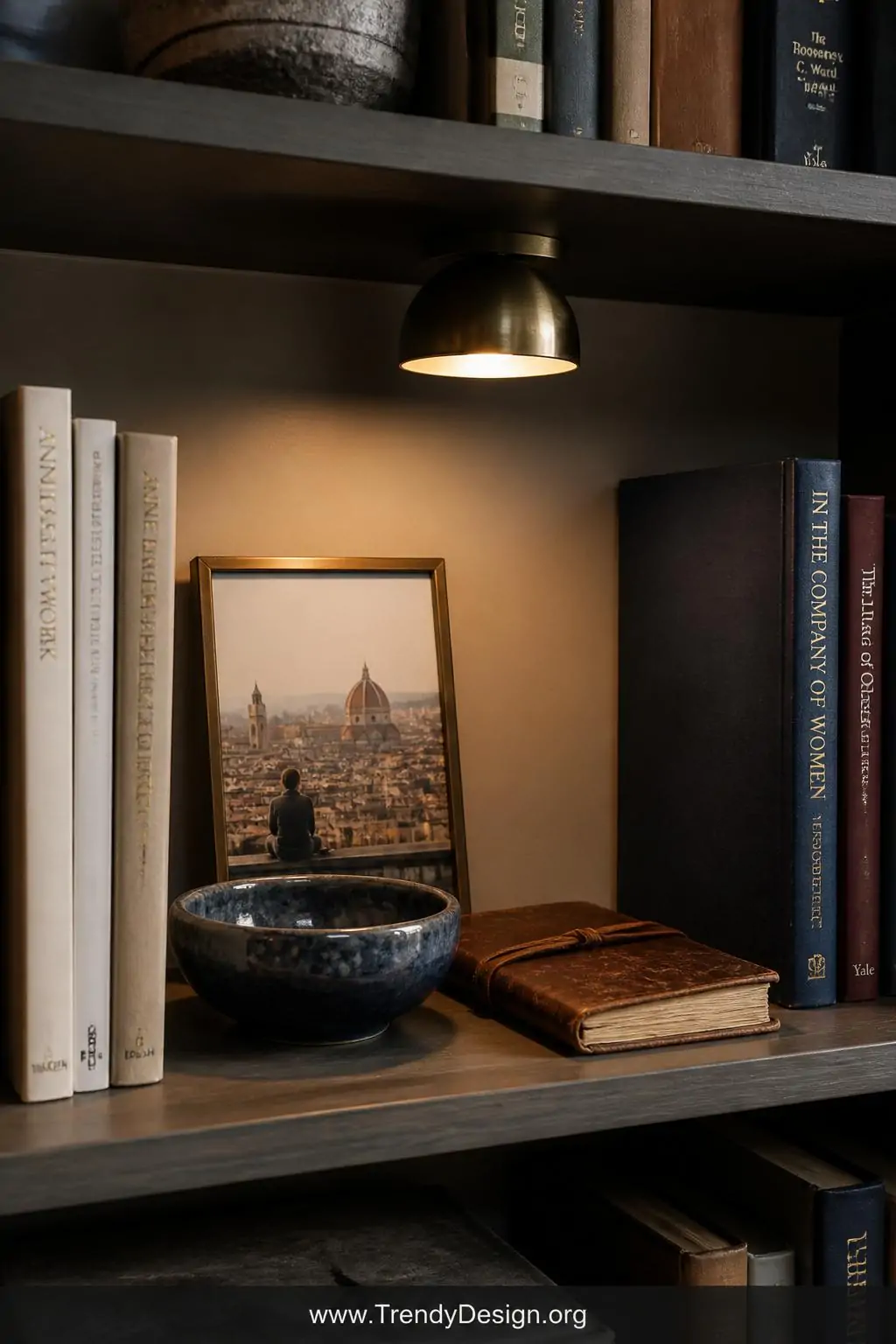 Medium shot of a styled bookshelf section where a small brass-framed travel photograph leans against the back wall beside a h