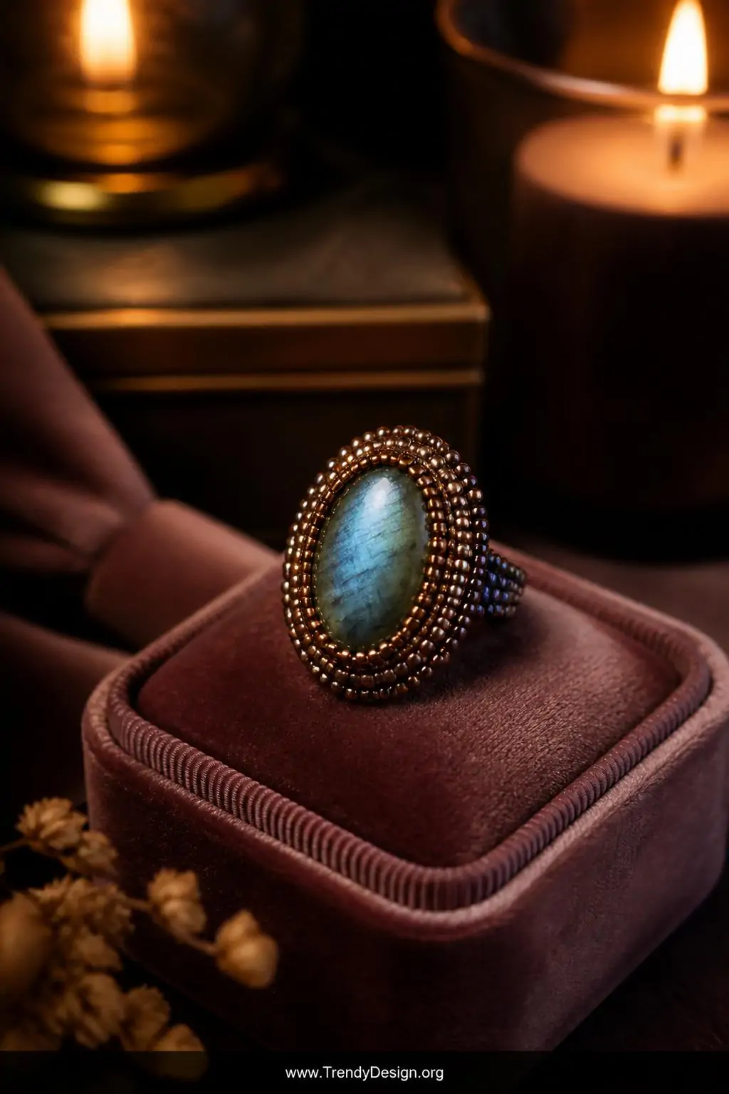 Beaded cabochon seed bead ring with a labradorite stone surrounded by iridescent gold seed beads displayed on a velvet ring cushion in dusty mauve, moody low warm candlelight, medium closeup emphasizing the jewel-like metallic bead bezeling