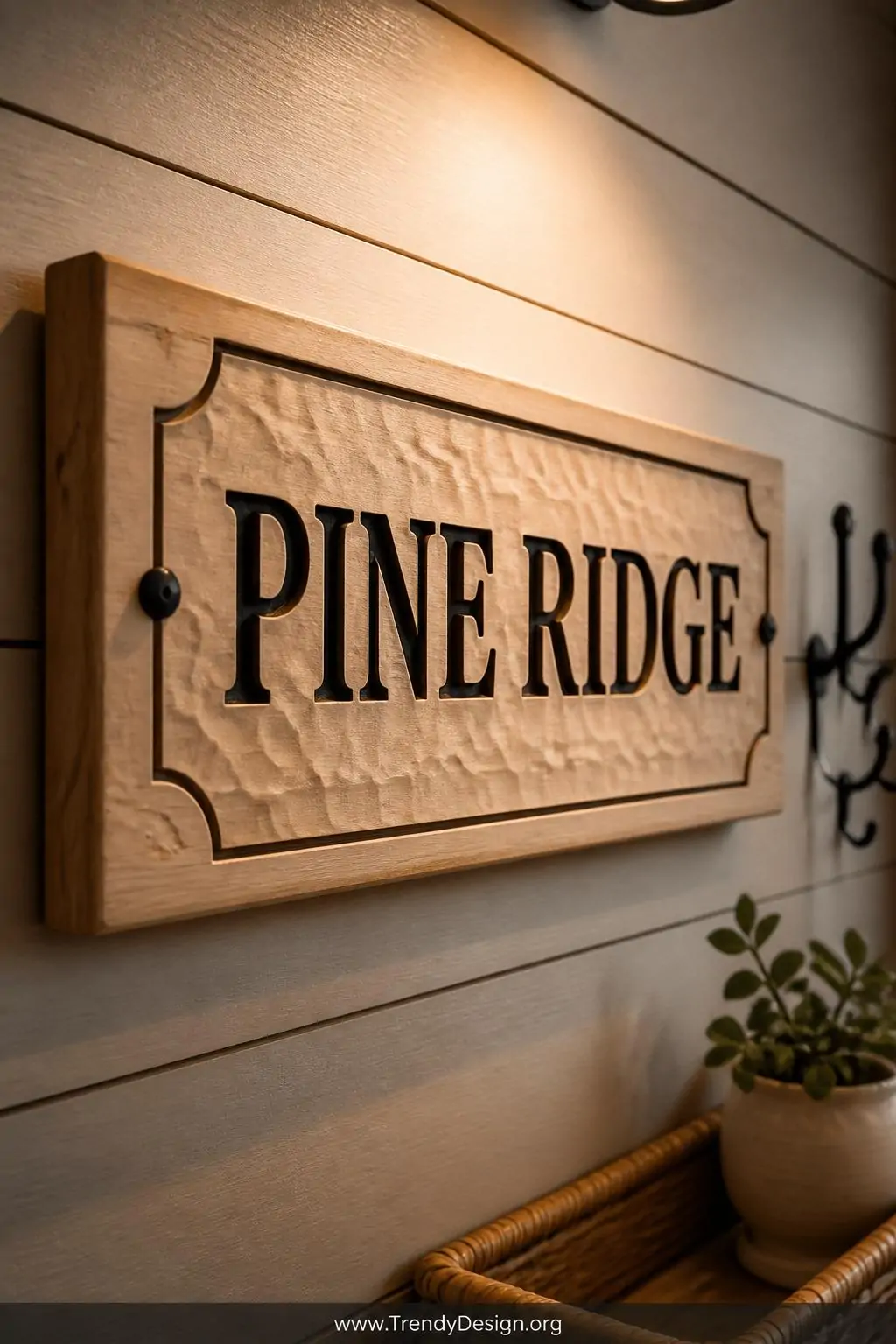 Carved wooden nameplate with deep chip-carved letters painted in matte black contrasting against the pale basswood grain, mounted on a shiplap wall in a warm neutral entryway, directional spotlight highlighting carved recesses, closeup shot