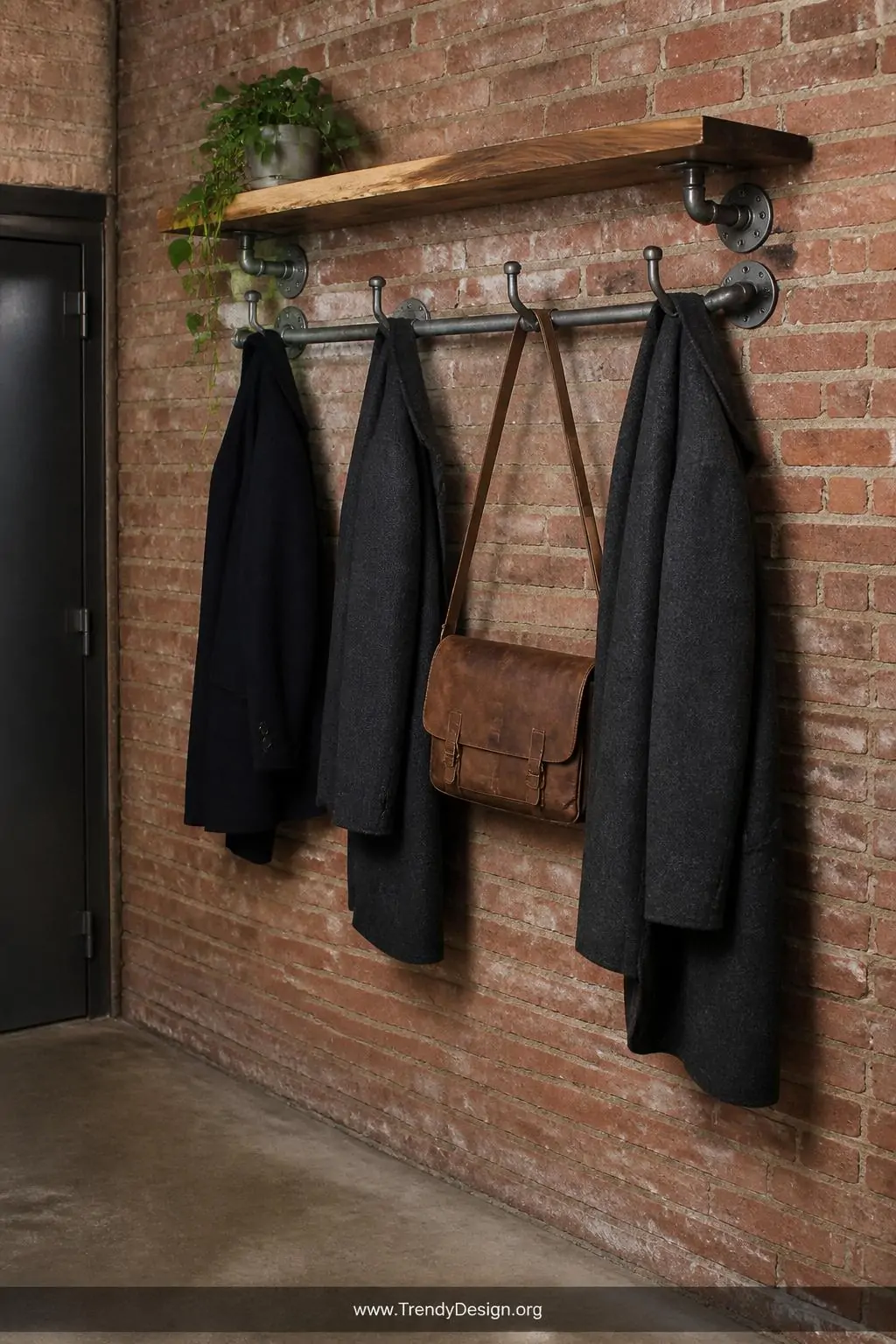 Galvanized steel pipe coat rack with a live-edge walnut shelf mounted above in an industrial entryway, raw pipe fittings and flanges visible against exposed brick wall, dark wool coats and a leather messenger bag hanging from pipe hooks, a small potted trailing plant on the upper shelf, concrete floor below, wide shot.