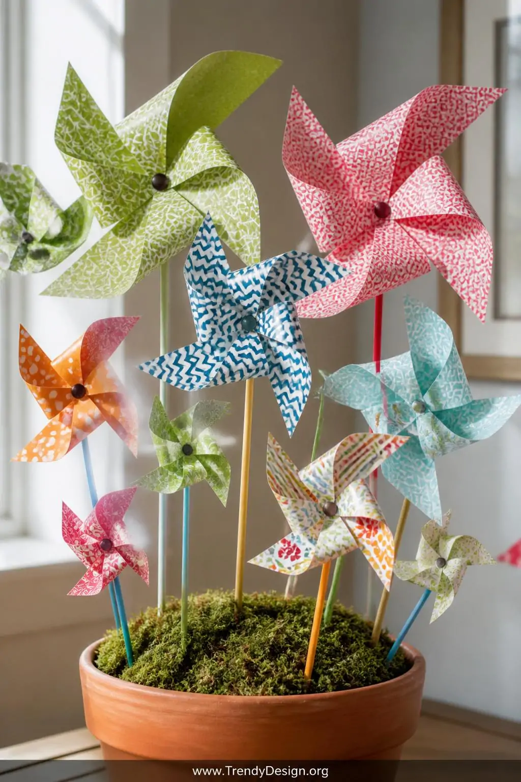 Paper pinwheel garden of mixed small and large patterned scrapbook paper pinwheels on colorful wooden skewers planted in a moss-covered foam block inside a terracotta pot, bright playful natural light, closeup shot emphasizing the geometric prints and layered color coordination.