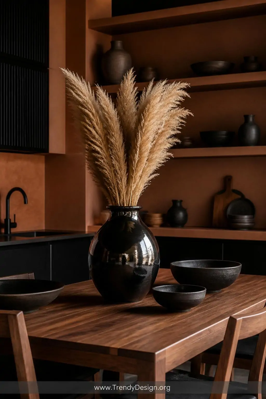 Jet-black barro negro ceramic vase with a mirror-polished metallic sheen holding dried pampas grass on a warm walnut dining table, surrounded by smaller black clay bowls on open terracotta-toned kitchen shelving, dramatic moody side lighting, medium shot of a sophisticated Mexican-modern kitchen.