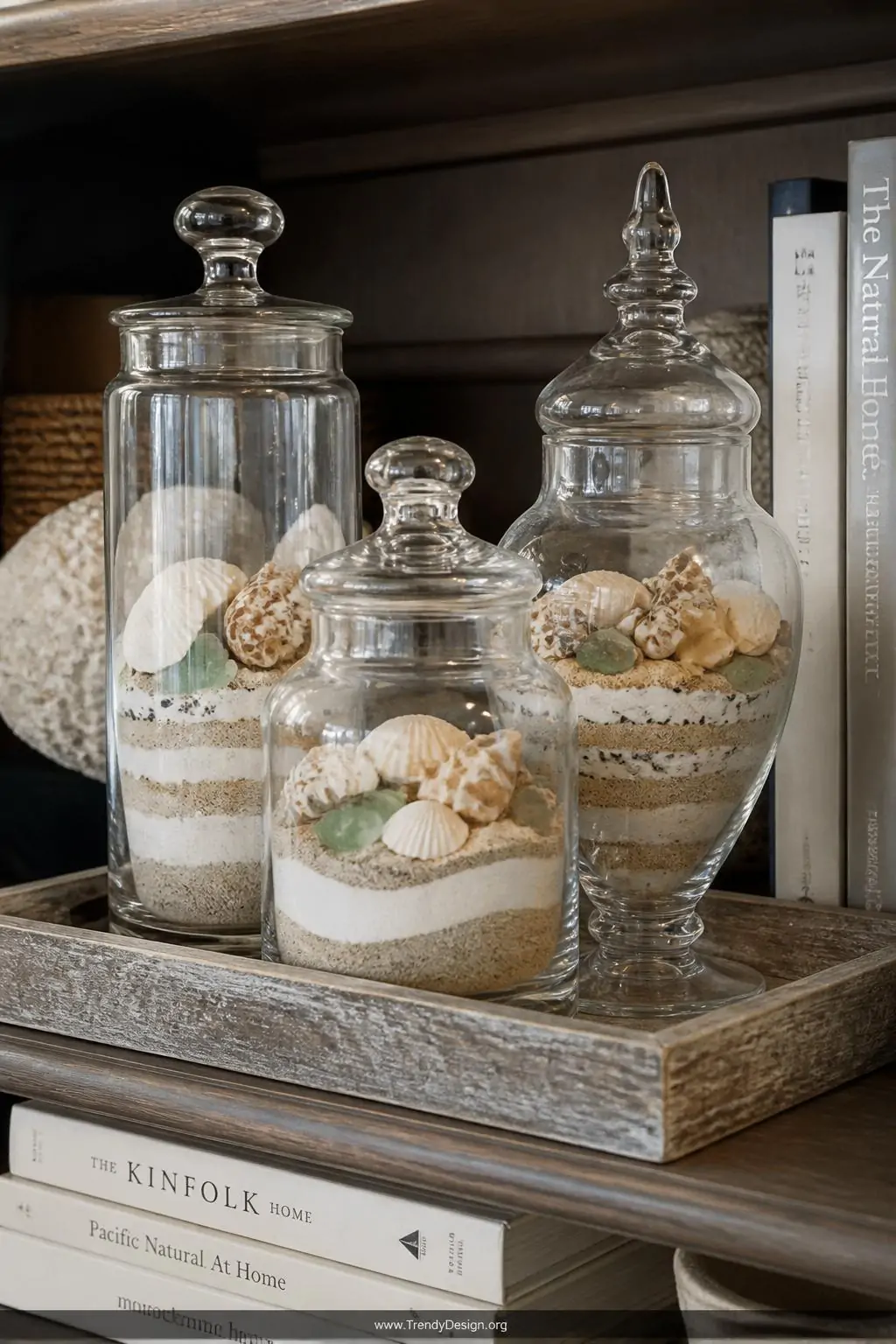 Three mismatched clear apothecary jars grouped on a weathered wood tray on a bookshelf, each filled with layered ivory and warm tan sand, embedded shells and beach glass, soft warm interior light, closeup
