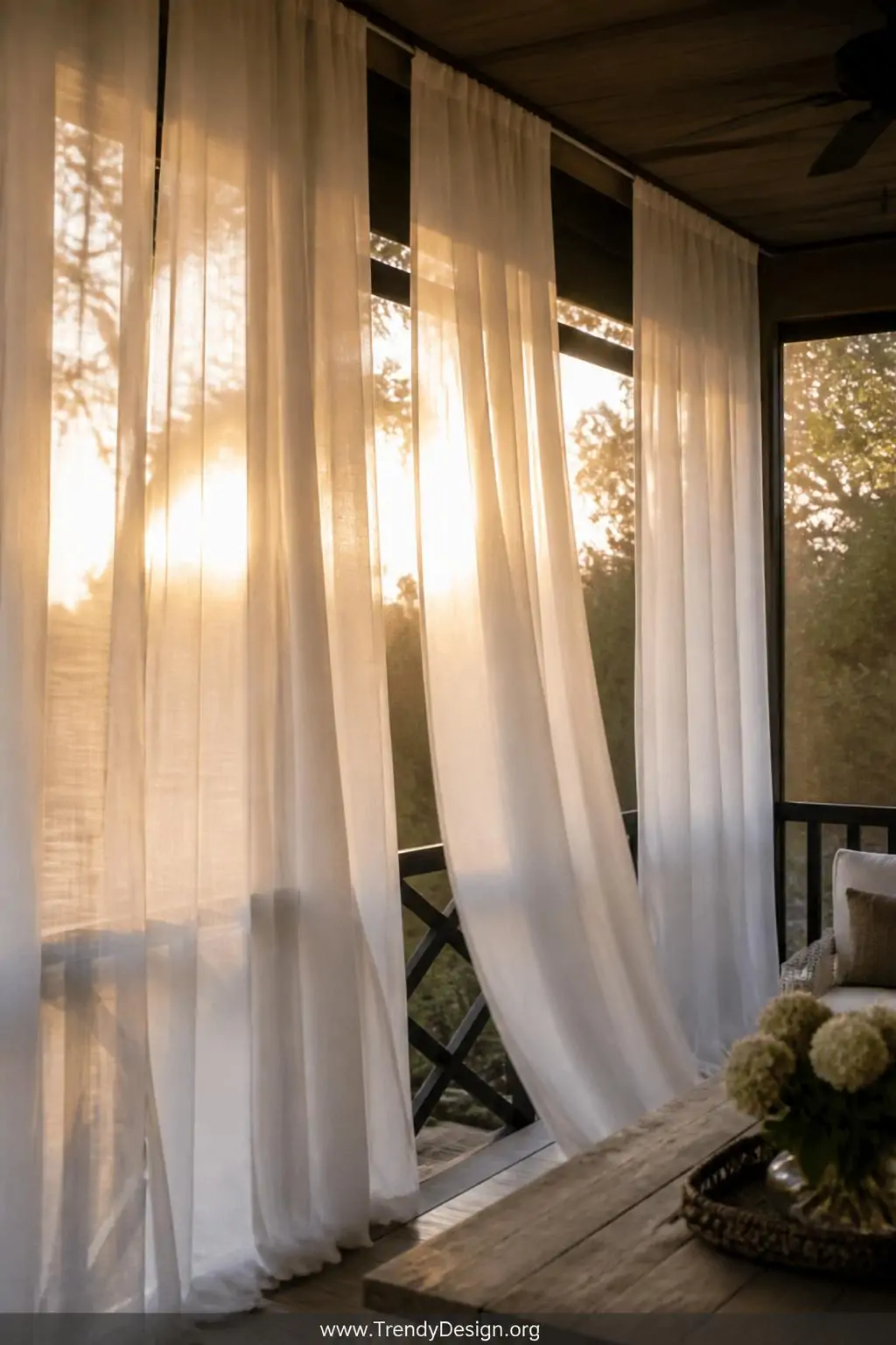 Sheer white linen outdoor curtain panels hanging floor-to-ceiling along screened porch walls, fabric billowing softly in a breeze against golden late afternoon light, medium shot capturing romantic layered texture and warm shadows.