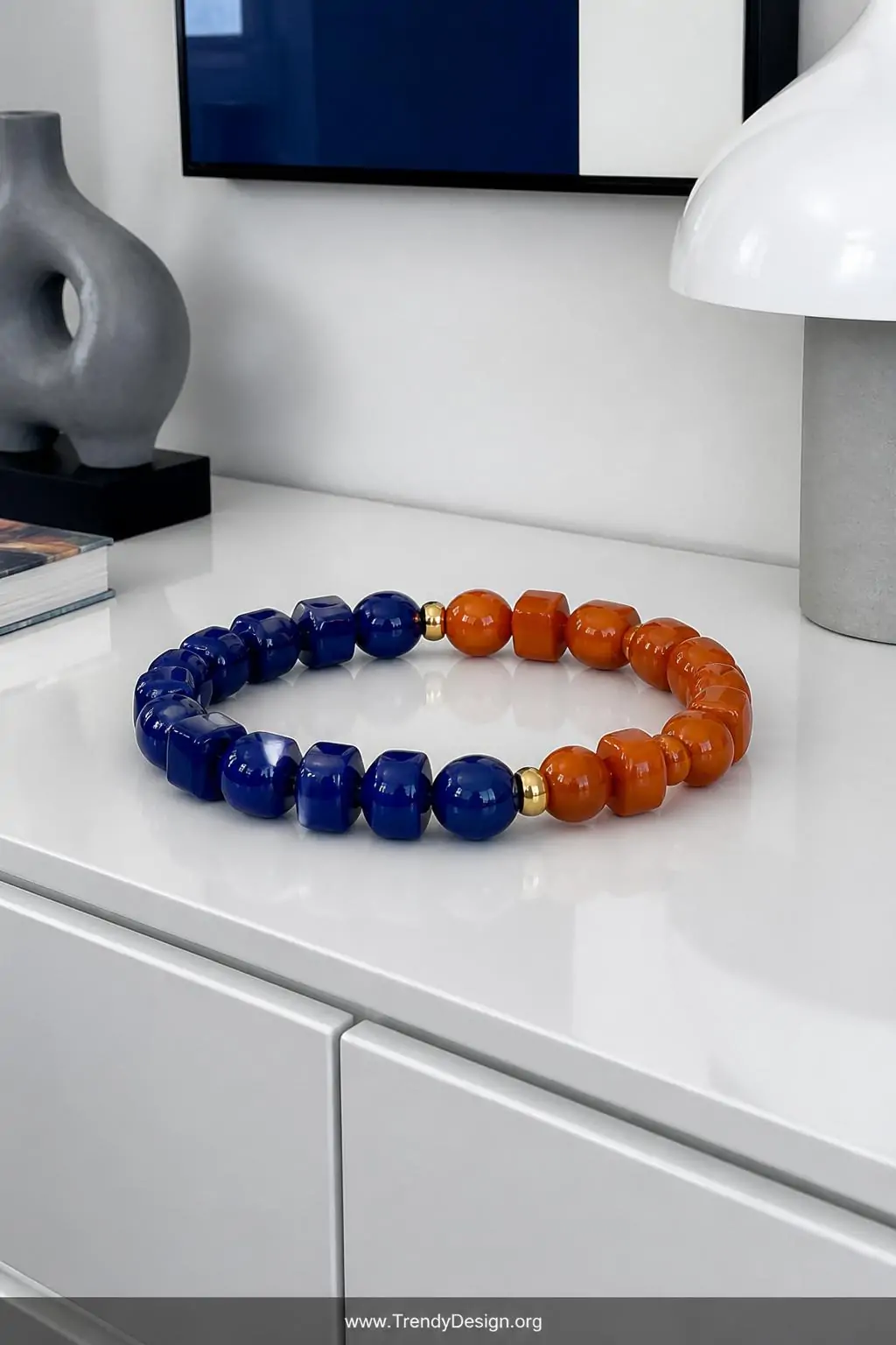 Bold color-blocked bracelet in distinct cobalt blue and burnt orange acrylic bead sections displayed flat on a sleek white lacquered console table, modern minimalist interior, medium shot with crisp cool overhead lighting emphasizing the sharp graphic contrast between the two vivid color sections.