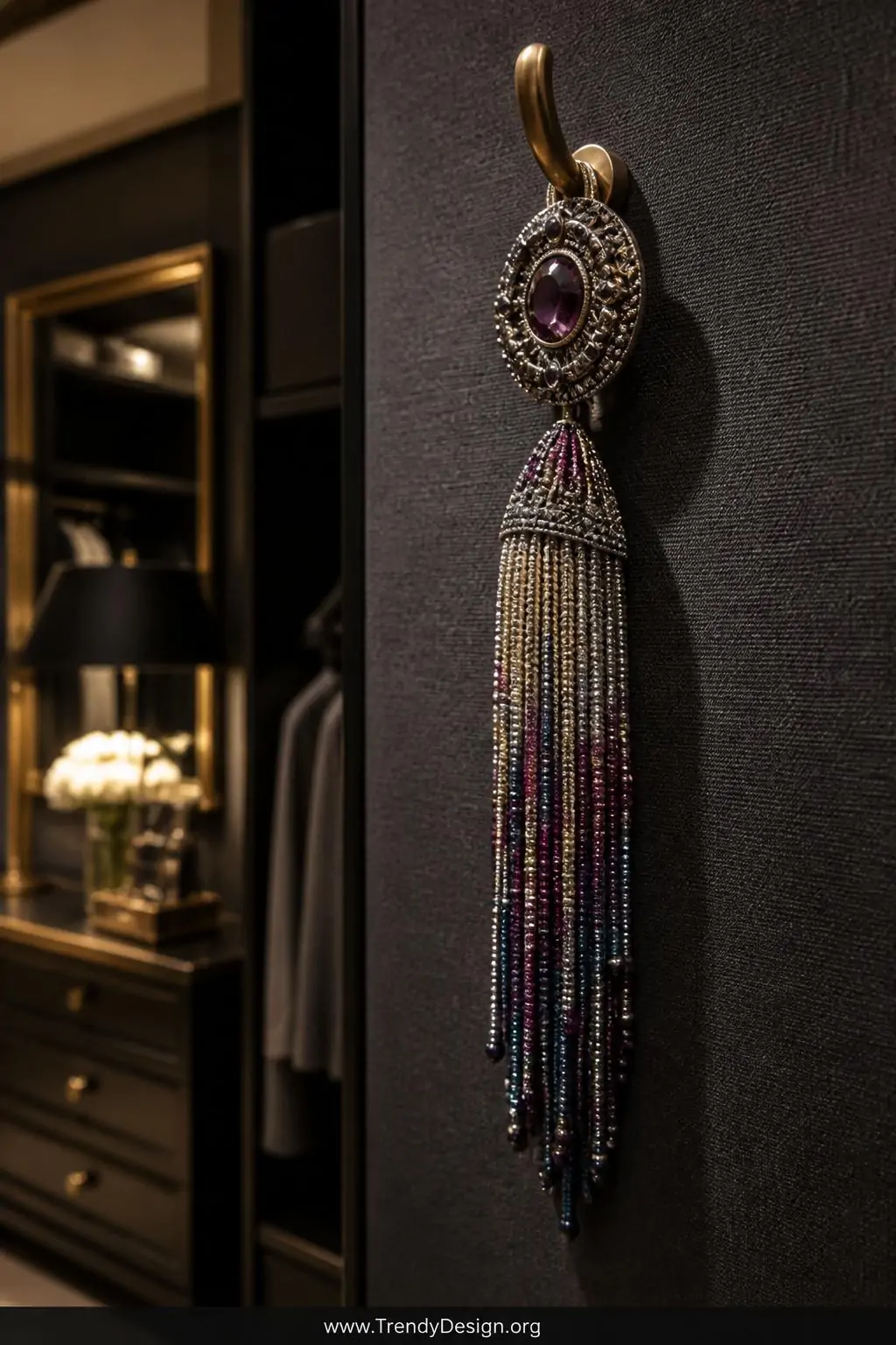 Beaded tassel brooch with cascading silver and jewel-toned seed bead fringe strands displayed hanging from a brass hook against a solid charcoal linen backdrop in an elegant dressing room, warm directional spotlight, closeup