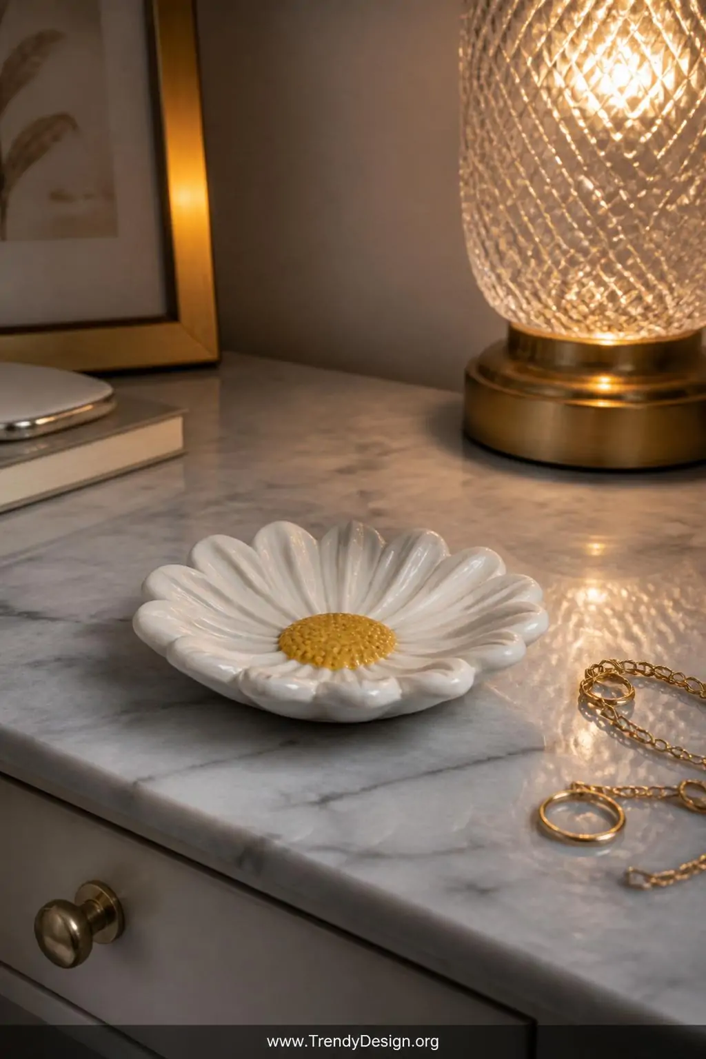 Clay daisy ring dish with white curling petals and bright yellow center sitting on a marble vanity nightstand, matte varnish finish catching warm lamp light beside delicate jewelry, closeup shot.