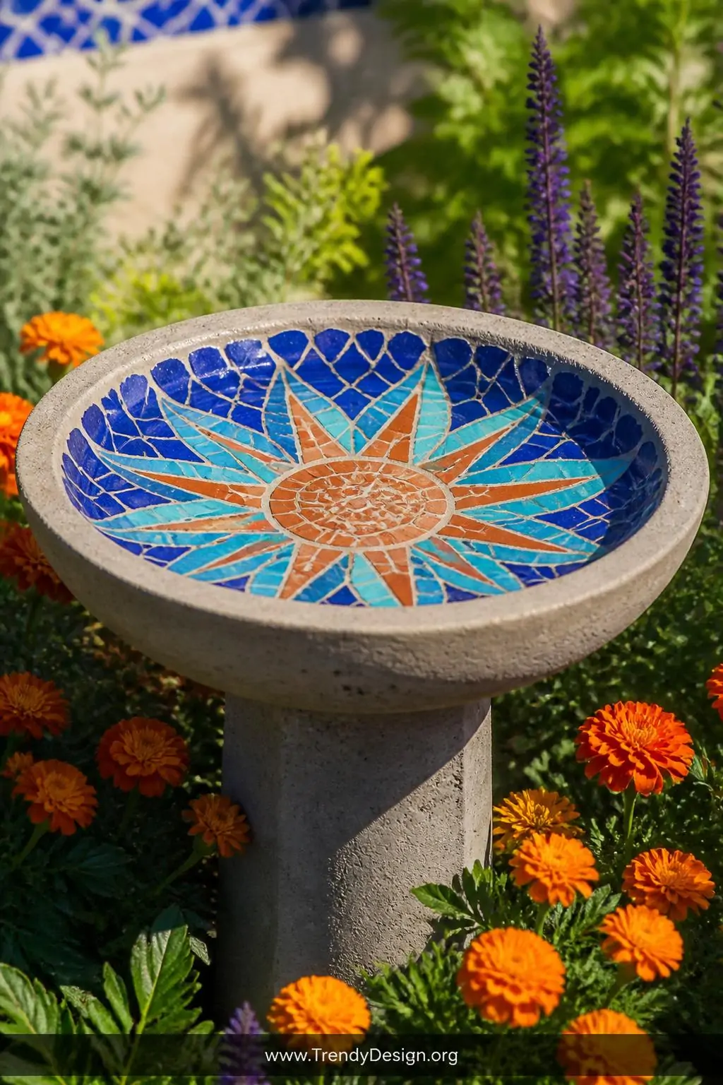 Mosaic-topped concrete bird bath bowl embedded with vibrant cobalt blue, turquoise, and terracotta broken ceramic tiles forming a geometric sunburst pattern, set on a simple concrete pedestal in a sun-drenched garden bed of bright marigolds and salvia, closeup shot.