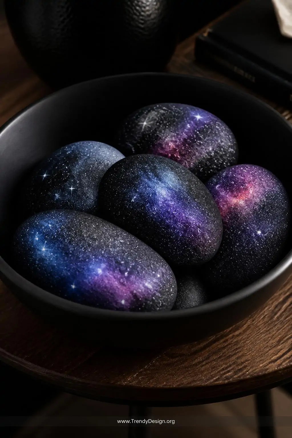 A closeup shot of galaxy-painted rocks resting inside a matte black ceramic bowl on a dark walnut side table, each stone feat