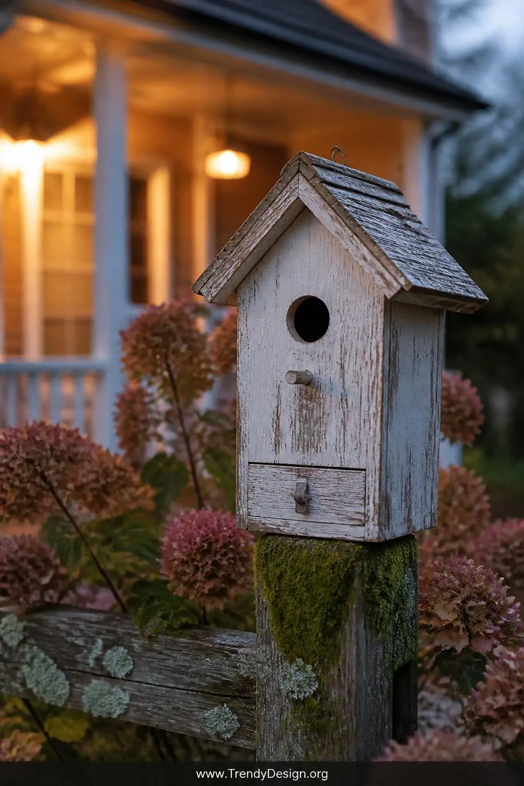 A closeup shot of a weathered whitewash wood birdhouse mounted on a mossy fence post at dusk, chipped cream paint revealing r