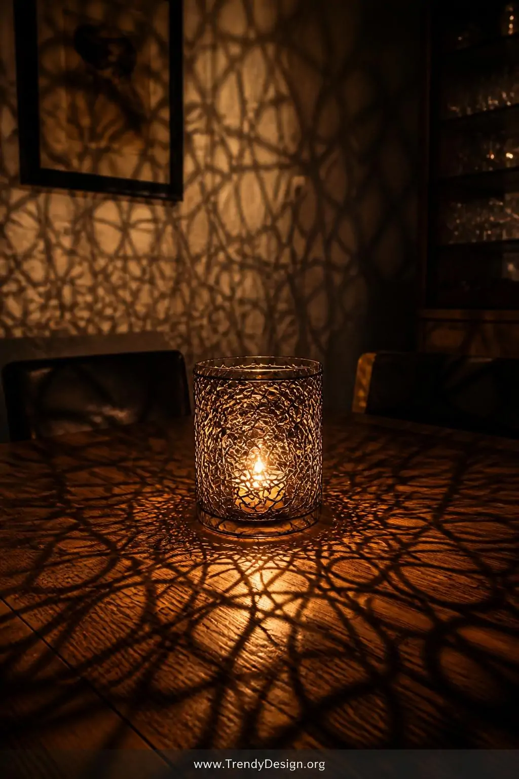 Coiled wire candle holder wrapped around a clear glass jar with a glowing amber tea light inside, intricate shadow lacework patterns radiating across a dark walnut dining table and surrounding walls, moody intimate dining room with no other light source, closeup shot capturing the warm golden glow and delicate wire shadow detail.
