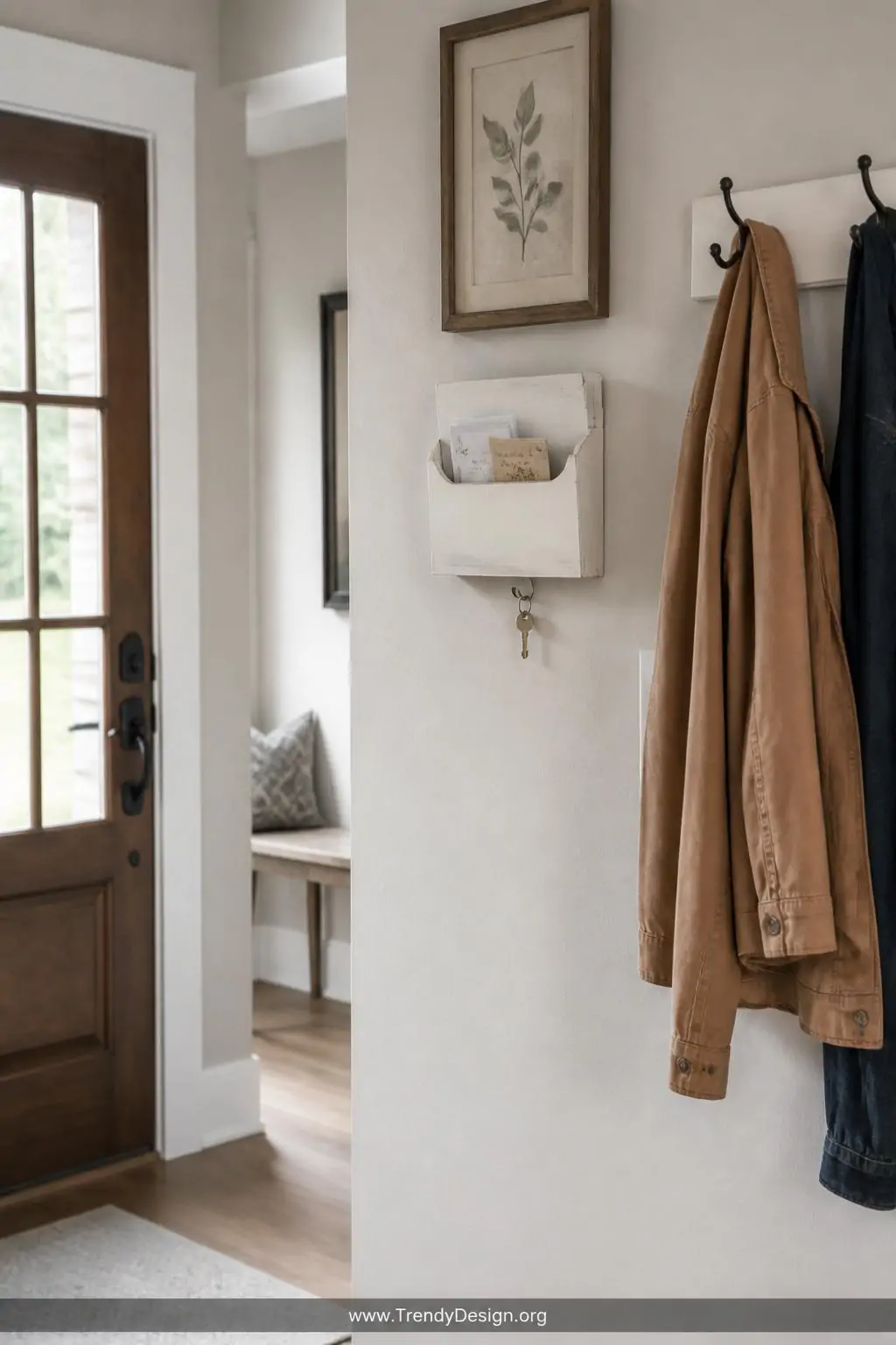 Shallow open-fronted wall-mounted wooden note box with white painted finish holding folded paper notes and a small key hook attached, installed on a light gray entryway wall beside a coat rack, bright natural hallway light, wide shot showing placement at eye level near the front door.