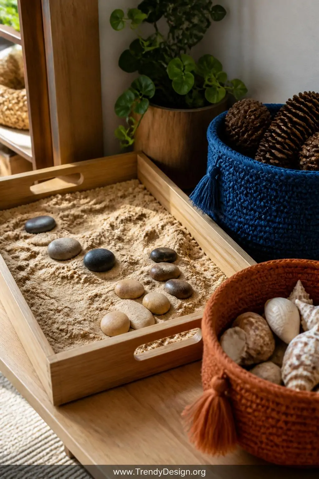 Wooden tray table holding kinetic sand and colorful river stones beside cobalt blue and terracotta woven baskets filled with pinecones and shells, closeup of a Montessori sensory discovery corner, soft natural side lighting emphasizing rich textures and earthy tones