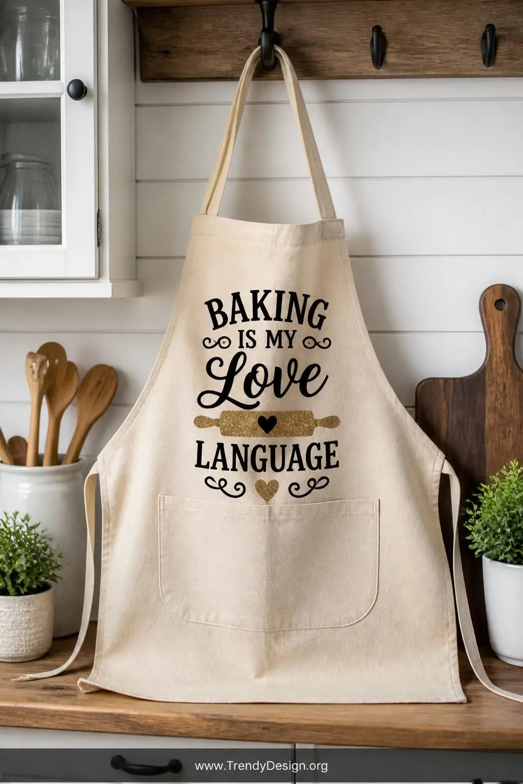 Canvas apron with centered matte black vinyl text and glitter accent food graphics hanging from a farmhouse kitchen hook, bright overhead lighting, medium shot highlighting the layered vinyl textures against the natural canvas weave.