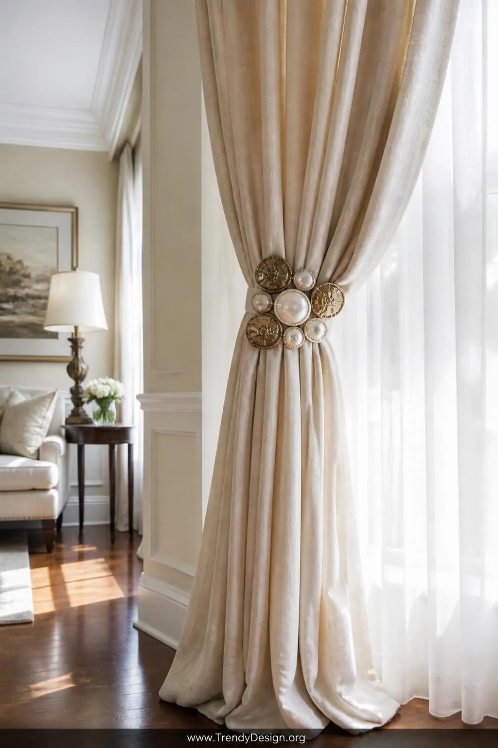 Fabric curtain tieback on ivory linen drapes clustered with a statement arrangement of oversized antique brass and pearl buttons at center, sun-drenched living room with hardwood floors and white trim, medium shot highlighting the window treatment detail against backlit sheer fabric.