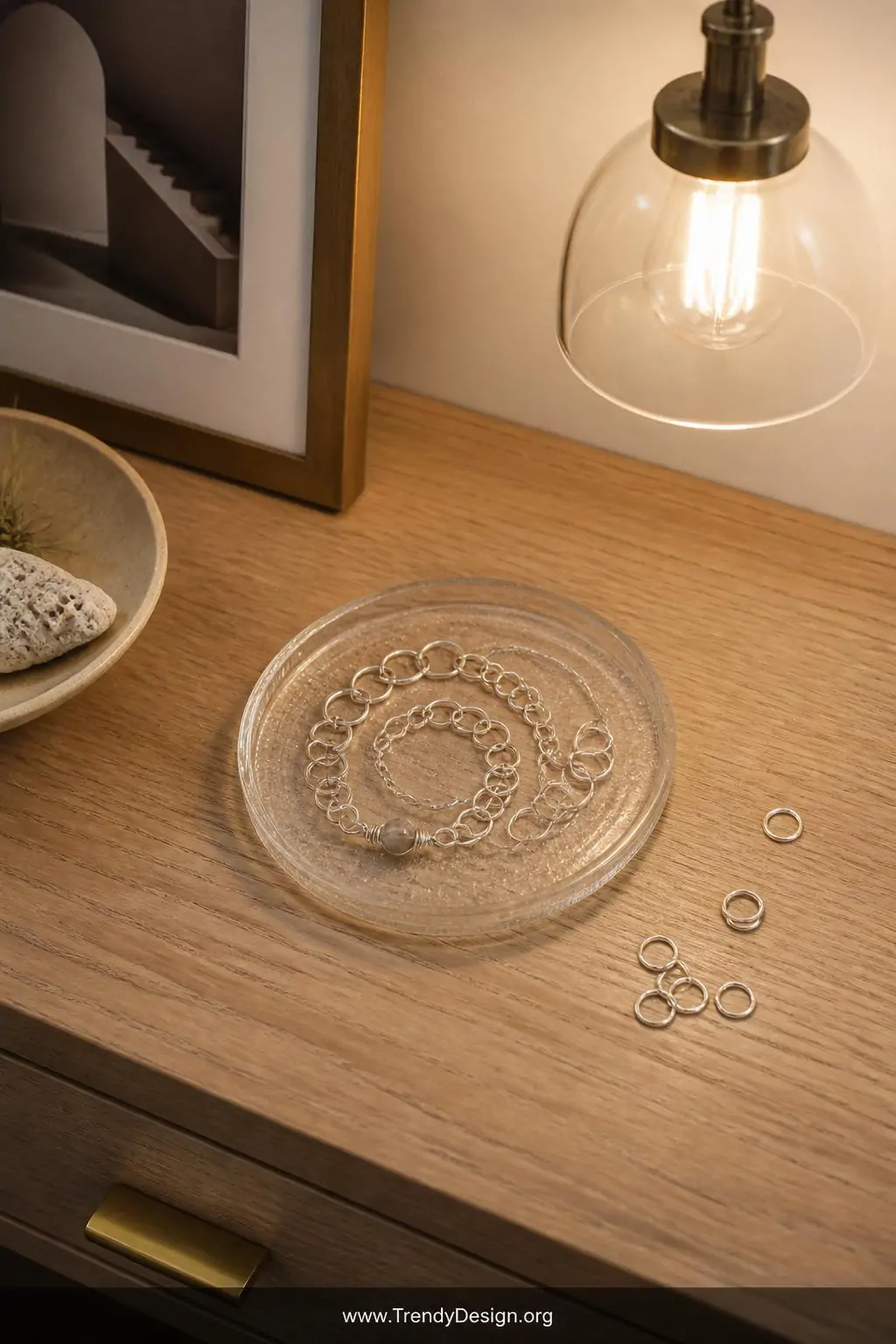 Adjustable wire ring-and-chain bracelet loosely coiled on a small glass jewelry dish beside scattered jump rings on a light oak desk, warm overhead pendant light, organic hand-formed silver links with a tiny wire-wrapped bead focal point, medium shot