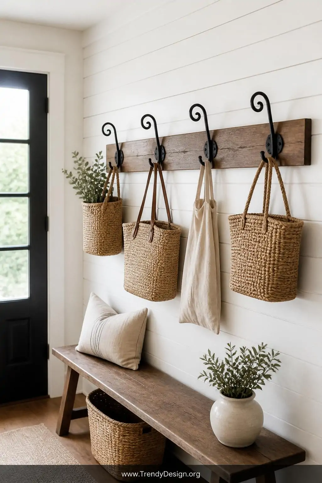 Set of four matching blacksmith-made iron scroll wall hooks mounted on a single stained oak backing board against crisp white shiplap, holding woven baskets and a linen tote in a bright farmhouse entryway under natural window light, wide shot.