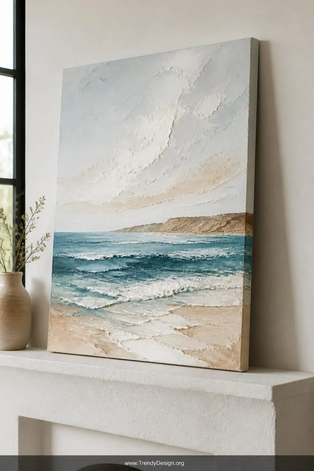 Textured acrylic canvas painting propped on a white plaster mantle, showing layered sandy ocean blue, tan, and white coastal seascape with visible three-dimensional palette knife strokes, bright natural window light, medium shot