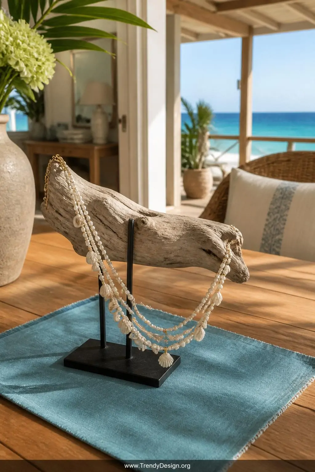 Three-strand layered shell and freshwater pearl anklet with gold seed beads cascading from a driftwood display stand on a sun-warmed teak outdoor table, turquoise linen napkin beneath, bright coastal afternoon light, wide shot, photorealistic interior design