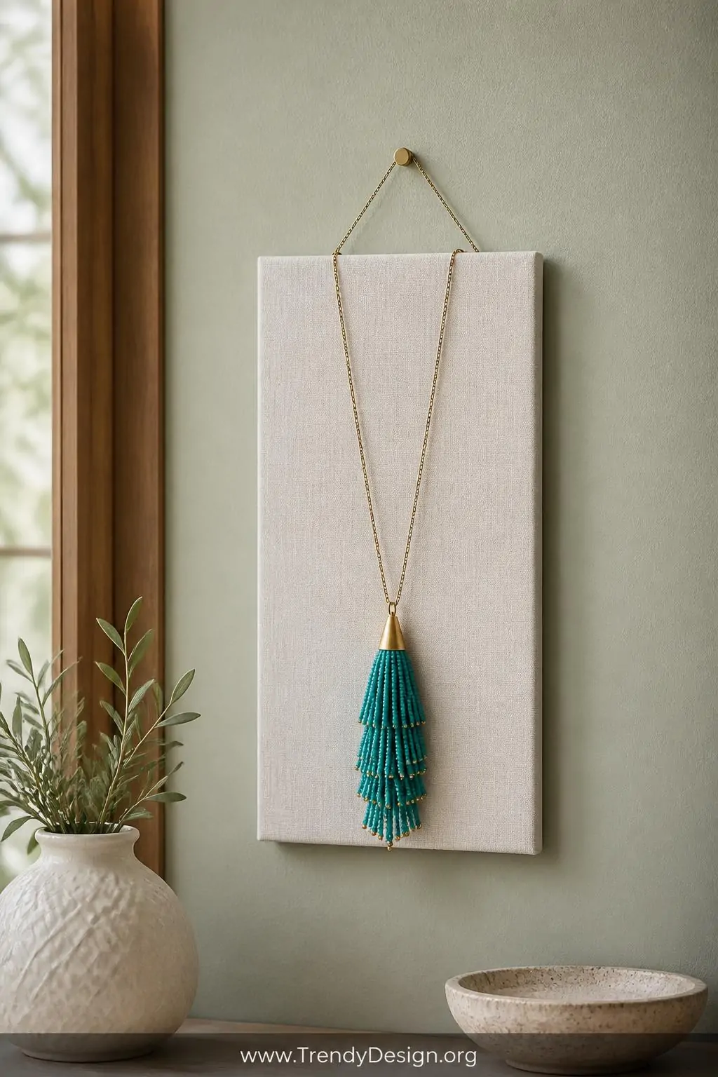 Turquoise seed bead tassel pendant necklace draped over a white linen textured display card hung on a soft sage green painted wall, bright diffused natural light, wide shot capturing the swishy layered pendant and delicate chain against the calm neutral background.