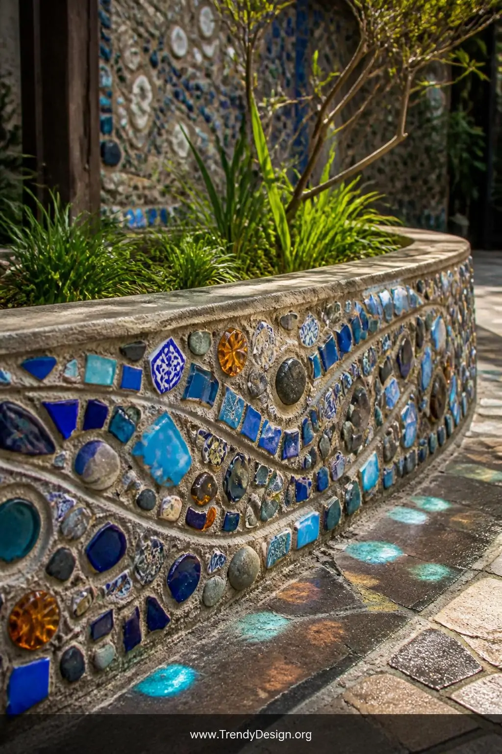 Mosaic-embedded concrete raised garden bed in a garden courtyard, artisan bohemian style, exterior walls encrusted with fragments of cobalt blue and turquoise ceramic tile, amber sea glass, and smooth river stones arranged in flowing geometric patterns, sunlight catching the glass pieces and scattering colored reflections onto surrounding pavers, closeup shot emphasizing the intricate embedded surface detail