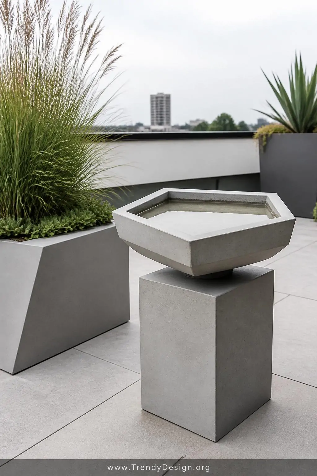 Minimalist geometric concrete bird bath with sharp angular hexagonal basin on a smooth cube pedestal, modern rooftop garden setting, contemporary architectural style, polished matte gray sealed concrete surface, paired with sleek angular planters and ornamental grasses, clean bright even daylight, wide shot emphasizing sculptural negative space and clean uncluttered surroundings.