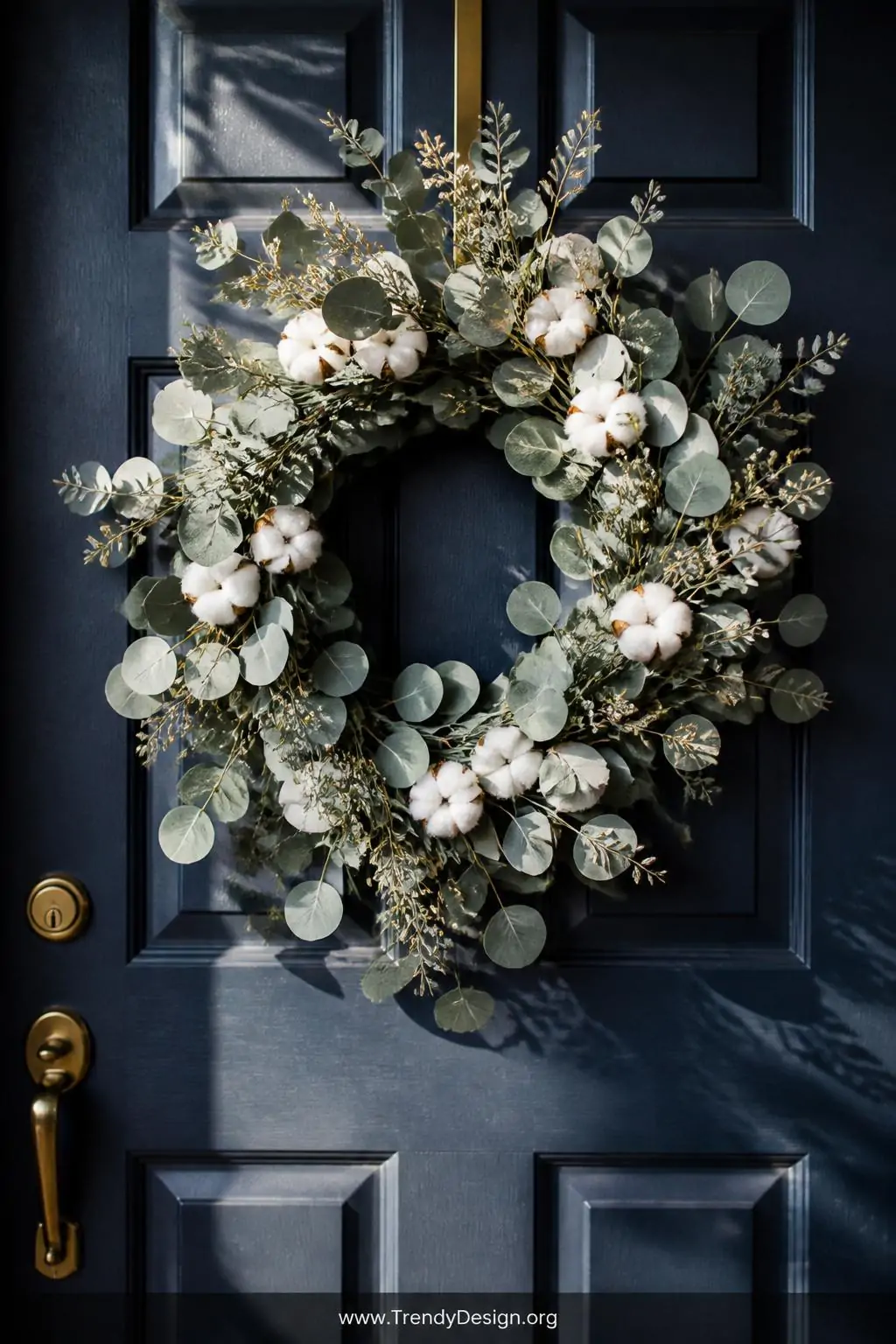 Closeup shot of a stunning year-round eucalyptus and dried cotton stem wreath hanging on a deep navy blue front door with bru