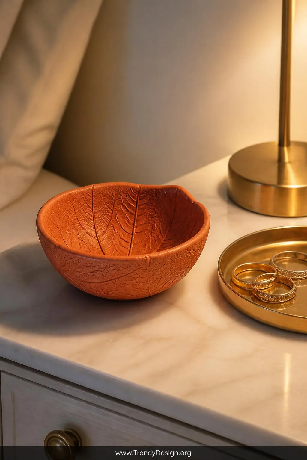 Small terracotta-painted air-dry clay bowl with deeply imprinted leaf vein texture resting on a marble dresser top beside a gold ring dish, warm bedroom lamp light casting soft shadows that highlight the botanical surface detail, closeup shot emphasizing the copper and clay tones.