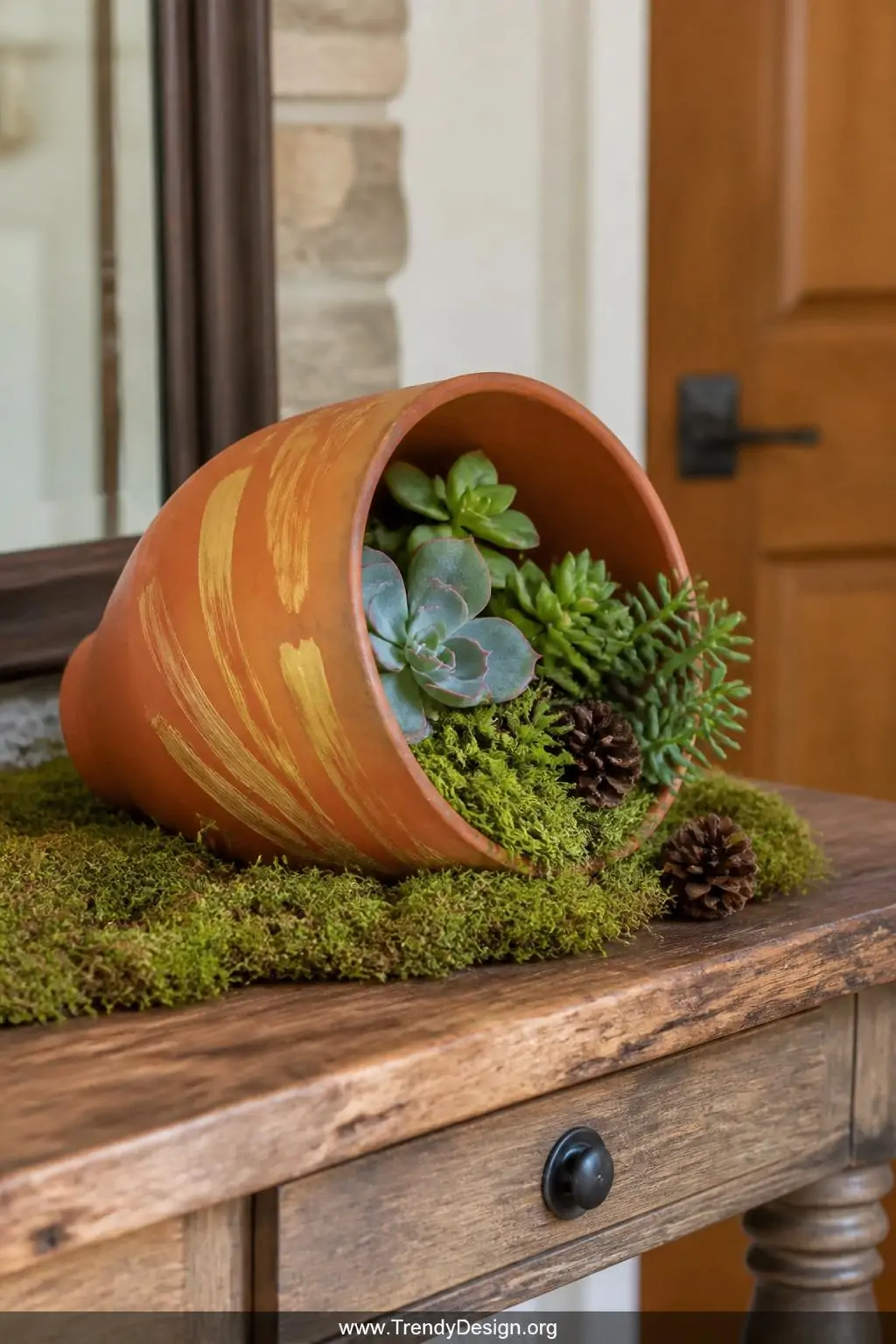 Terracotta pot painted in burnt orange and golden yellow brushstrokes lying sideways on faux moss, filled with succulents and mini pine cones on a rustic wooden entry table, closeup, photorealistic interior design