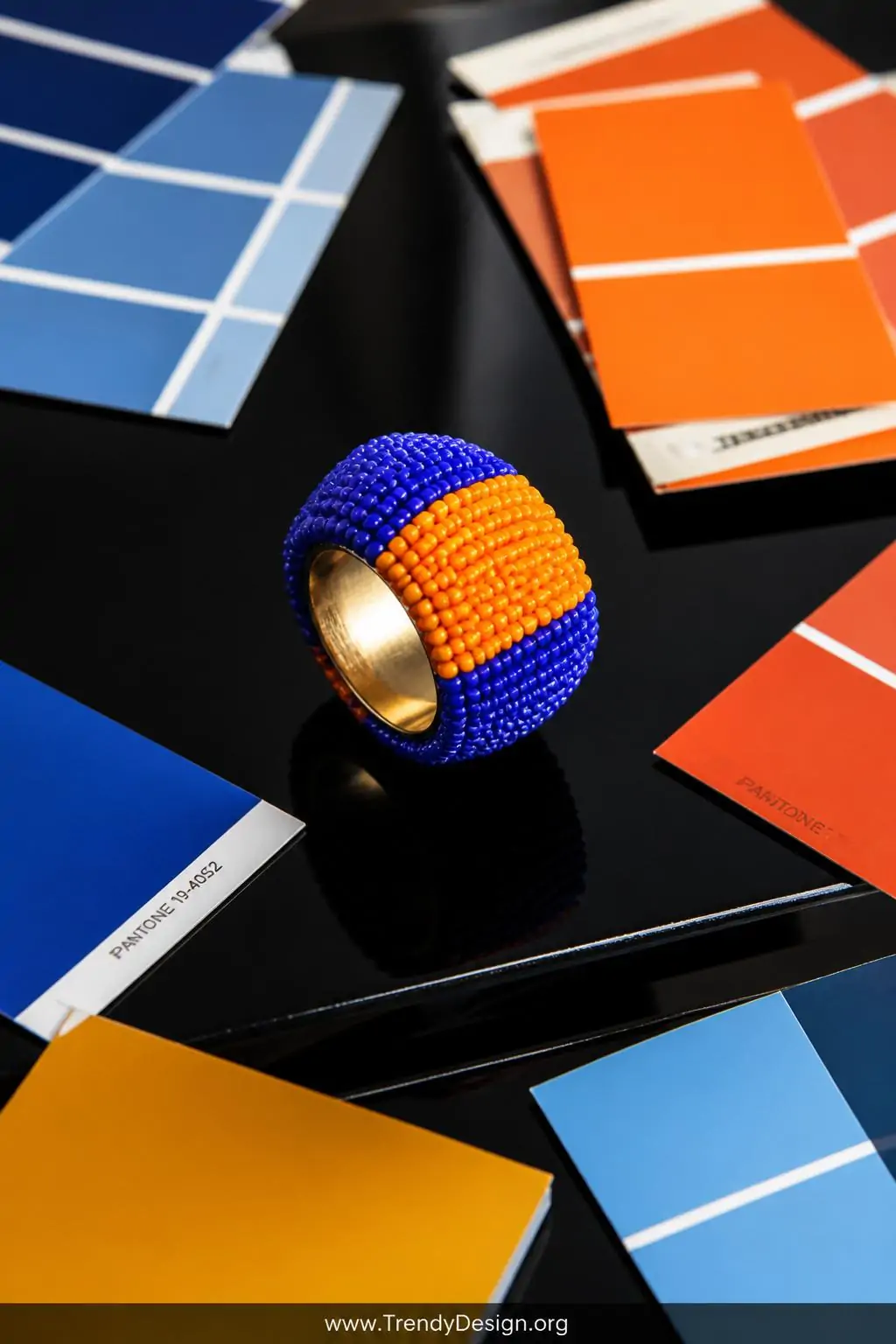 Wide color-blocked seed bead statement ring in royal blue and bright orange bands displayed dramatically on a glossy black lacquered surface beside scattered bold paint chip swatches, strong overhead studio light, wide shot with vivid graphic color impact