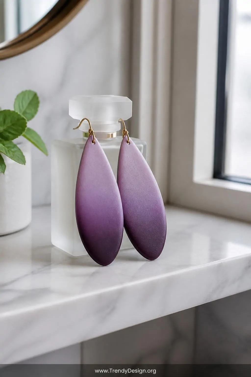 Gradient ombre polymer clay earrings blending dusty lilac into deep plum in elongated teardrop shapes propped against a small frosted glass perfume bottle on a marble bathroom shelf, cool north-facing window light revealing the seamless color transition across each earring surface, closeup shot.
