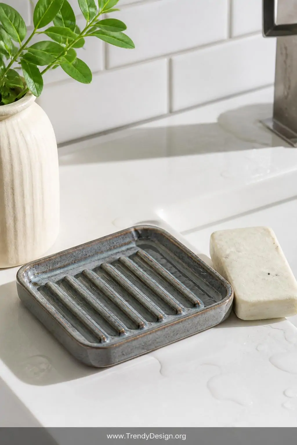 Handmade clay soap dish with parallel raised drainage ridges in a cool matte glaze sitting beside a bar of soap on a wet white ceramic sink surround, bright bathroom light, closeup shot
