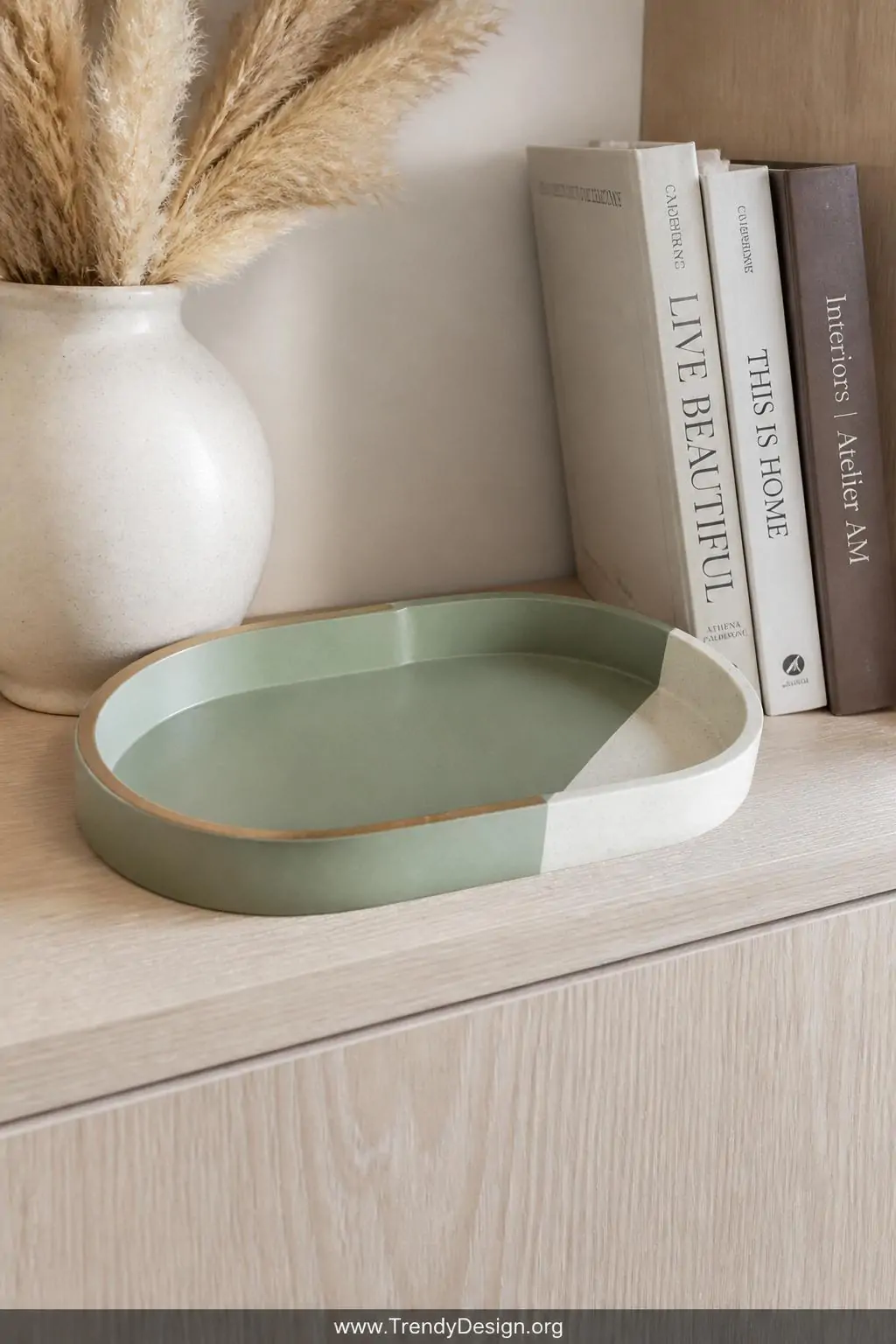 DIY painted concrete tray with crisp color-blocked sage green and creamy white edges and a delicate gold rim, resting on a white oak shelf beside dried pampas grass and a stack of design books, bright natural light, closeup shot.