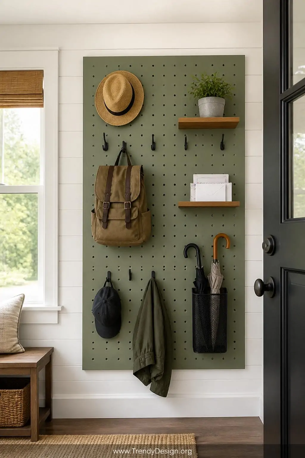 Sage green painted pegboard wall panel in a small entryway fully customized with matte black metal hooks holding hats and a canvas backpack, two small mounted wooden shelves holding a potted herb and a set of stacked mail, a metal mesh bin holding rolled umbrellas, styled cohesively against a white shiplap wall with natural light from a nearby window, wide shot, photorealistic interior design.