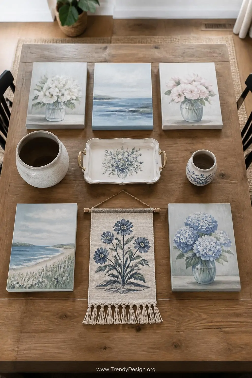 Flat lay of all eight finished student art projects arranged on a wide rustic farmhouse dining table including canvases, pots, tray, and wall hanging, bright overhead natural light, wide shot