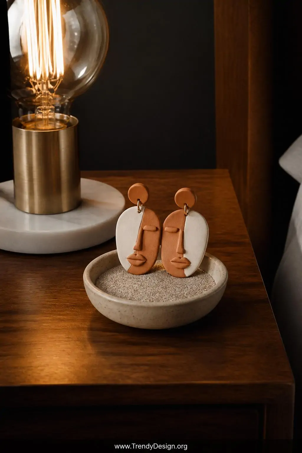 Sculptural miniature face polymer clay earrings in terracotta and ivory with tiny abstract nose and lips details displayed upright in a small sand-filled ceramic bowl on a mid-century modern nightstand, warm Edison bulb lamp casting dramatic side shadows that highlight the dimensional sculpted portrait features, closeup shot.
