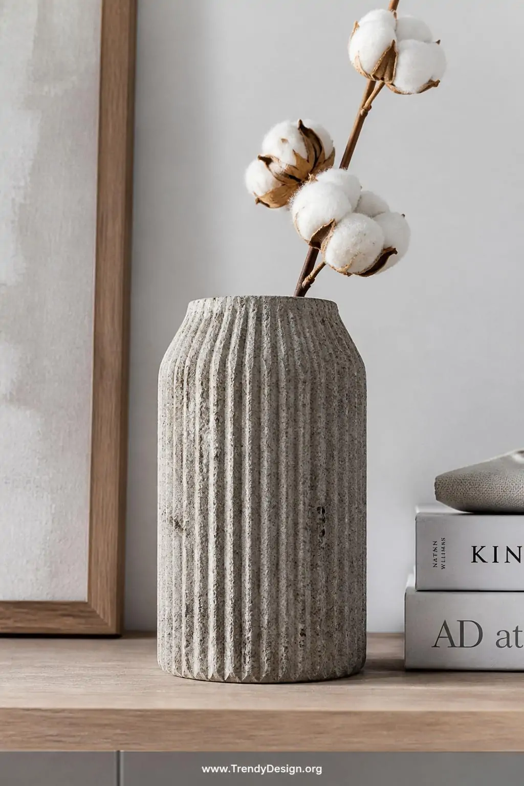 Textured ridged concrete vase with embedded pebble surface holding a single cotton flower stem on a minimalist wooden shelf, cool gray and natural tones, crisp daylight highlighting every surface variation, simple white backdrop, closeup shot, photorealistic interior design, no people.