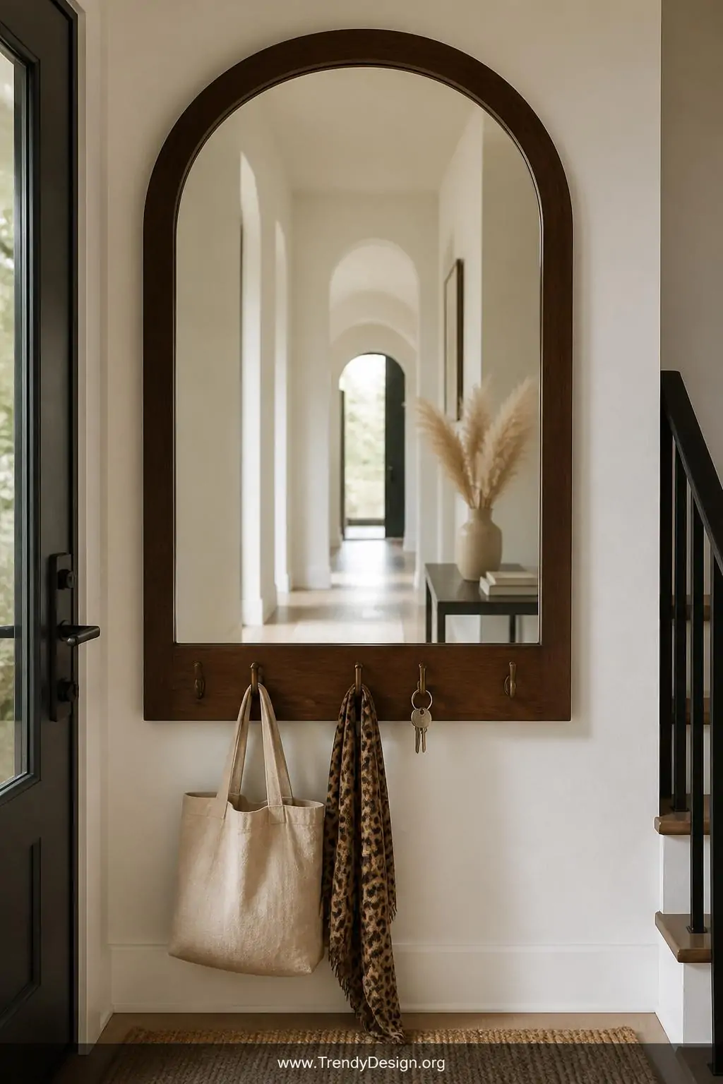Full-length arched mirror with a dark walnut frame lining the entryway wall, a row of five brushed bronze hooks along the bottom edge suspending a linen tote bag, a leopard print scarf, and a set of keys, the mirror reflecting a bright airy hallway and a small bouquet of dried pampas grass on a side table, medium shot, photorealistic interior design.