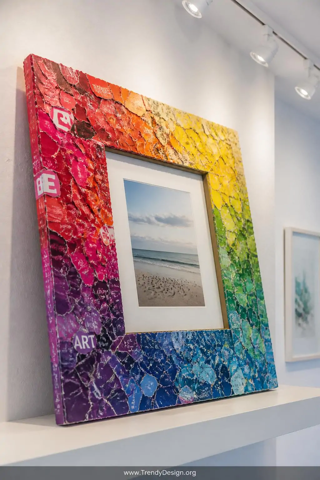 Mosaic magazine piece covered wooden picture frame in a vibrant rainbow tonal arrangement surrounding a displayed photograph, leaning against a white plaster wall on a floating shelf, bright contemporary gallery space, overhead track lighting illuminating every torn paper edge and color transition in sharp detail, closeup shot.