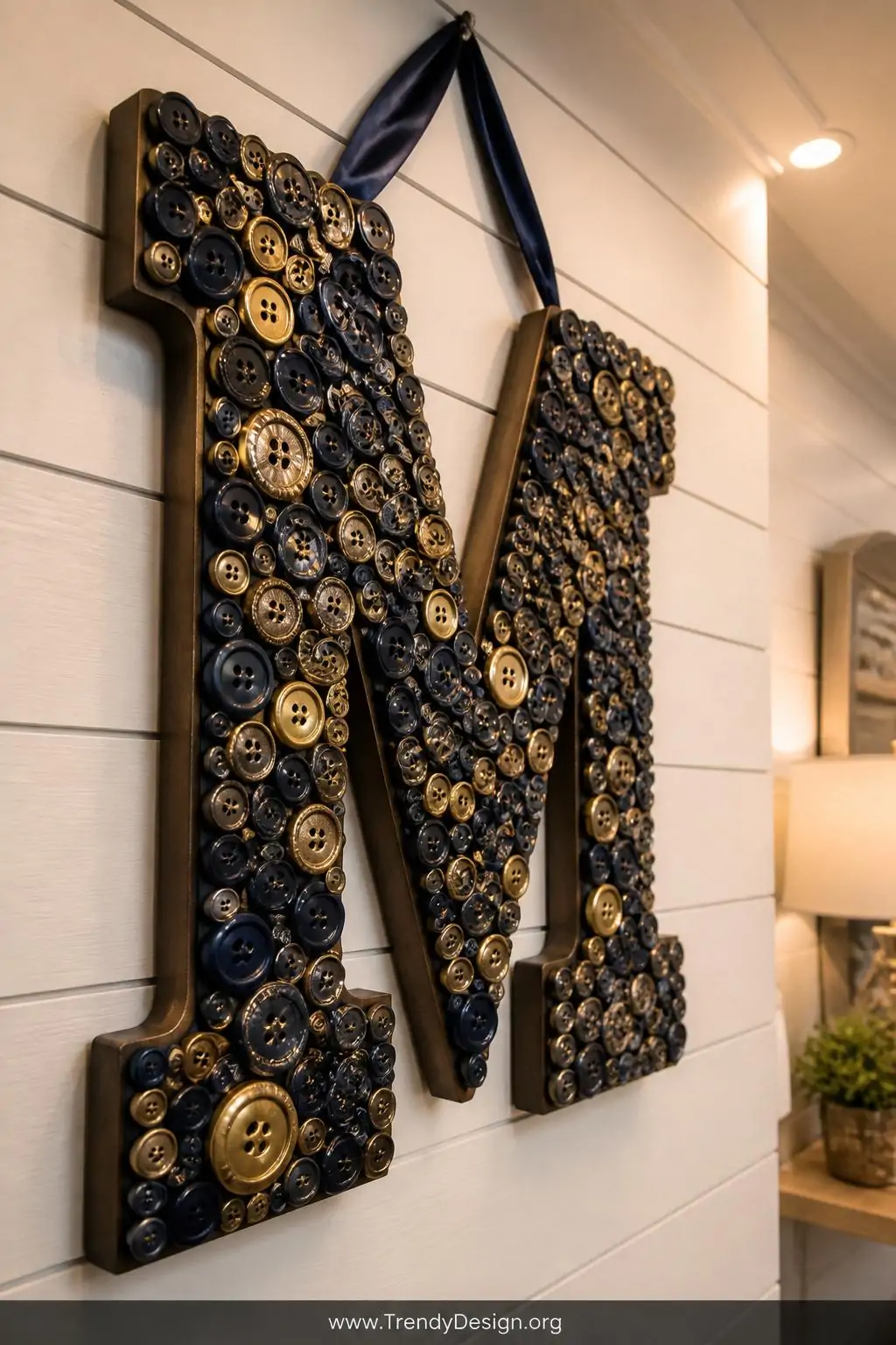 Large wooden letter M mounted on a shiplap wall completely covered in stacked buttons across a navy and gold color palette with varying depths and sizes, hanging by a satin ribbon loop, warm recessed lighting casting soft shadows, closeup shot emphasizing the three-dimensional handcrafted texture.