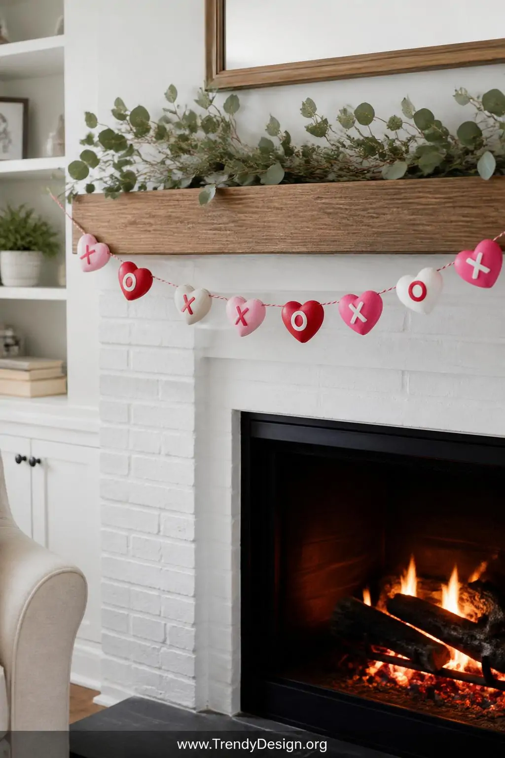 Colorful salt dough heart garland in red pink and white with painted X's and O's strung across a white brick fireplace mantle with eucalyptus sprigs, warm glowing firelight, wide shot.