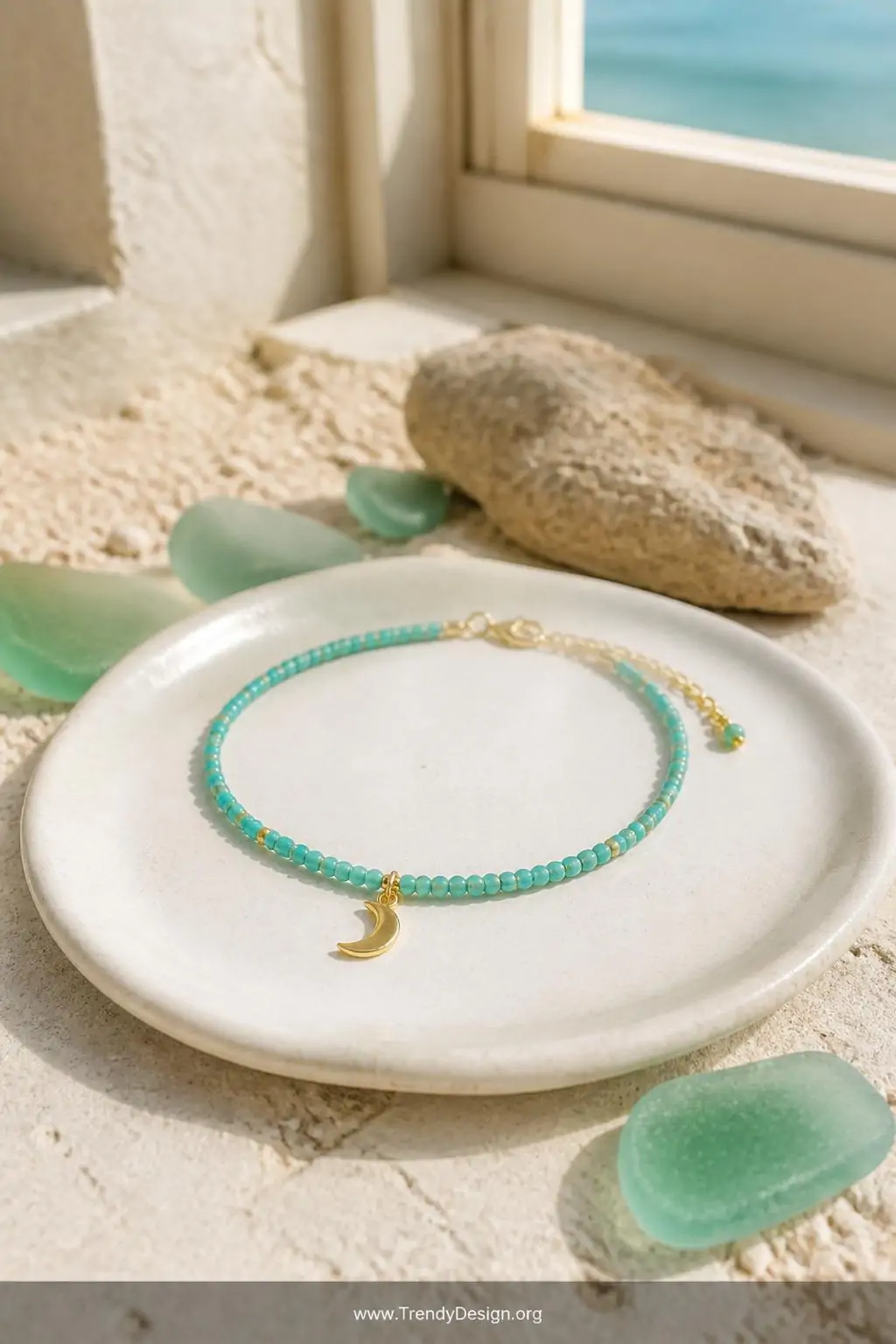 Delicate turquoise bead anklet with tiny gold seed bead accents and a small moon charm displayed on a smooth white ceramic dish near a sun-drenched window with sandy textures and sea glass nearby, bright warm light, closeup showcasing the ocean-inspired color palette and sparkling charm detail.