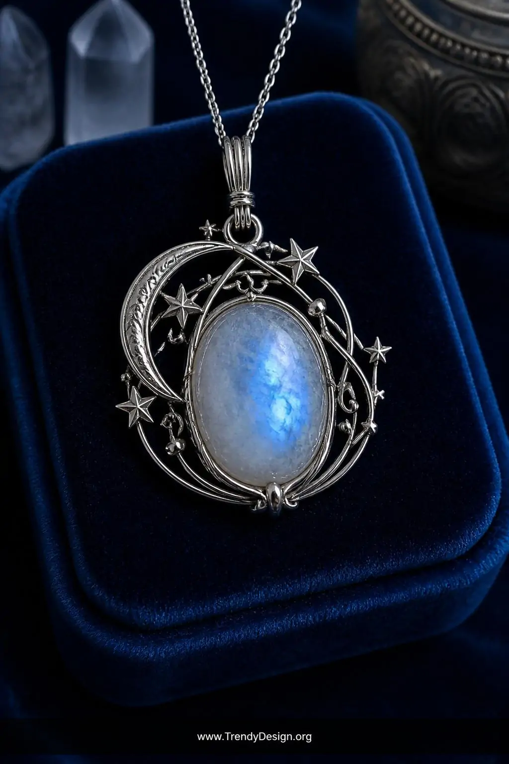 Oval moonstone pendant surrounded by sculpted 26-gauge fine silver wire stars and crescent moon details displayed on a navy blue velvet ring cushion, cool blue-toned studio light amplifying the stone's ethereal adularescence glow, medium closeup showing radiating celestial wire details