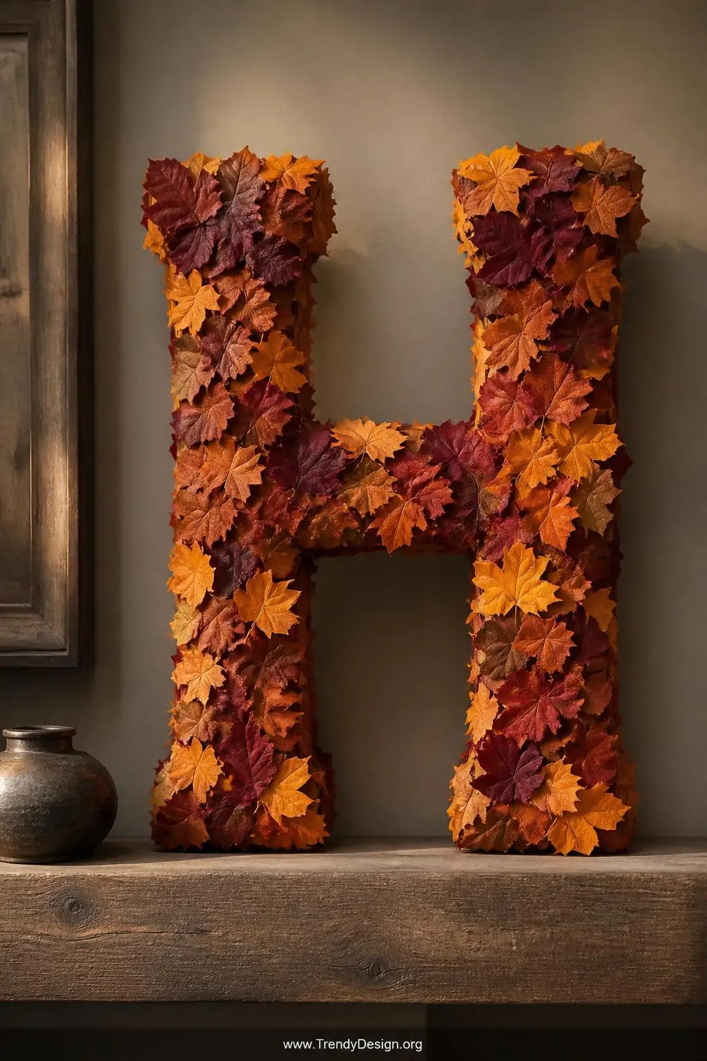 Large wooden letter H completely covered in overlapping real leaves in deep burgundy, burnt orange, and golden yellow tones secured with hot glue, propped on a reclaimed wood shelf against a warm greige accent wall with soft directional spotlighting, closeup shot highlighting the rich layered texture.