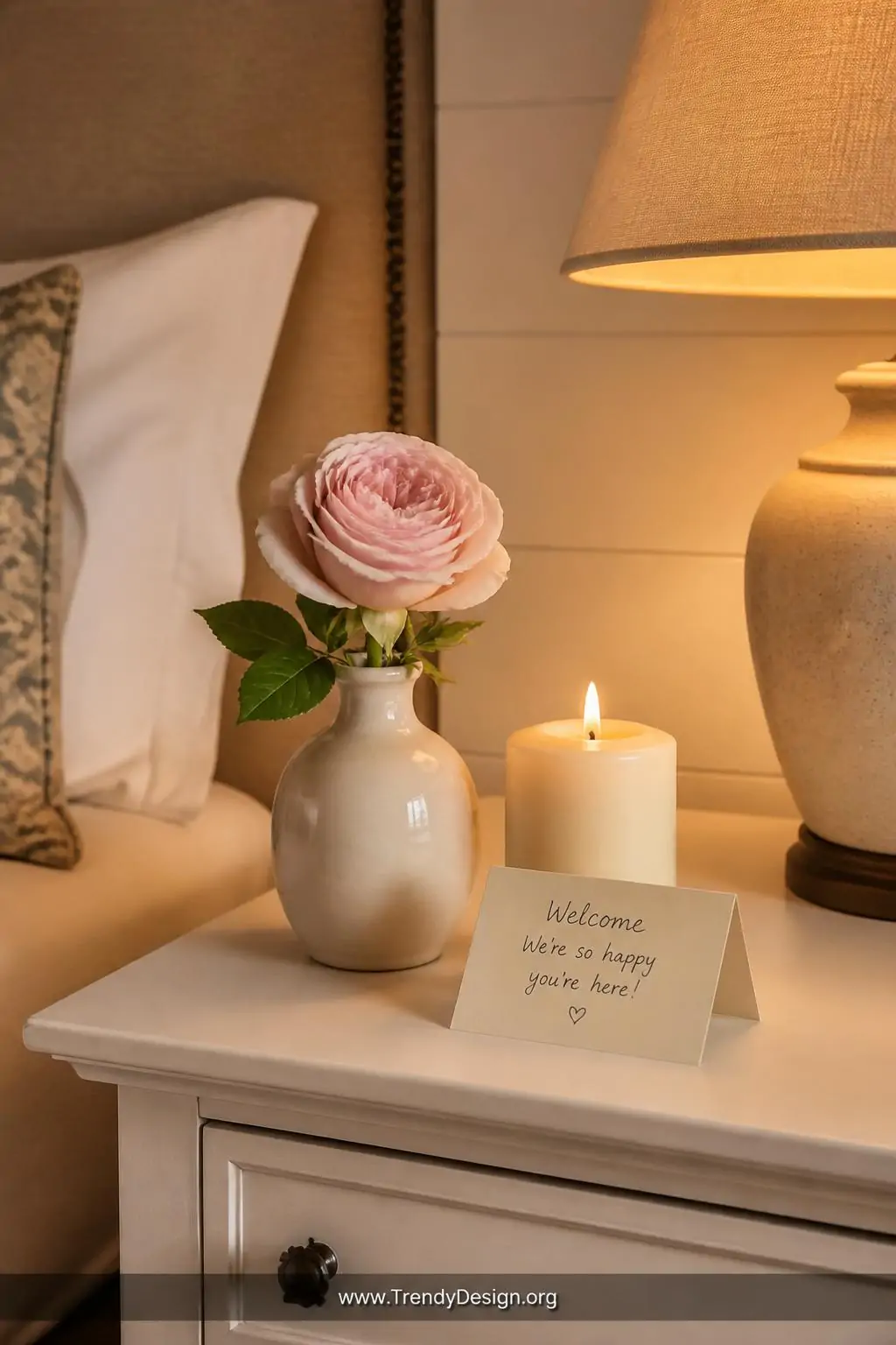 Single cream ceramic bud vase with a fresh pale pink garden rose beside a lit vanilla pillar candle and a small folded handwritten welcome note on a white wooden nightstand, warm golden lamp glow, intimate closeup shot, photorealistic interior design.