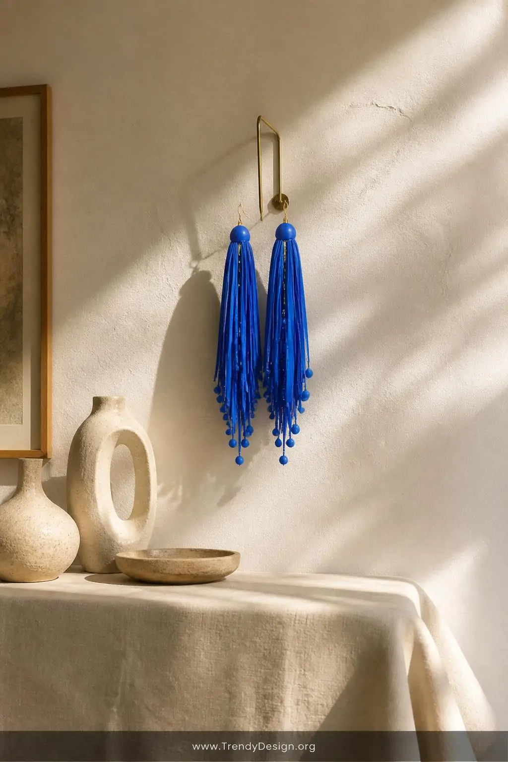 Statement fringe polymer clay earrings with long layered clay strips in bold cobalt blue mixed with small clay bead drops hanging from a slim brass wall hook against a warm white textured plaster wall above a linen-draped console table, wide shot with raking afternoon sunlight catching the movement and dimension of every cascading strip.