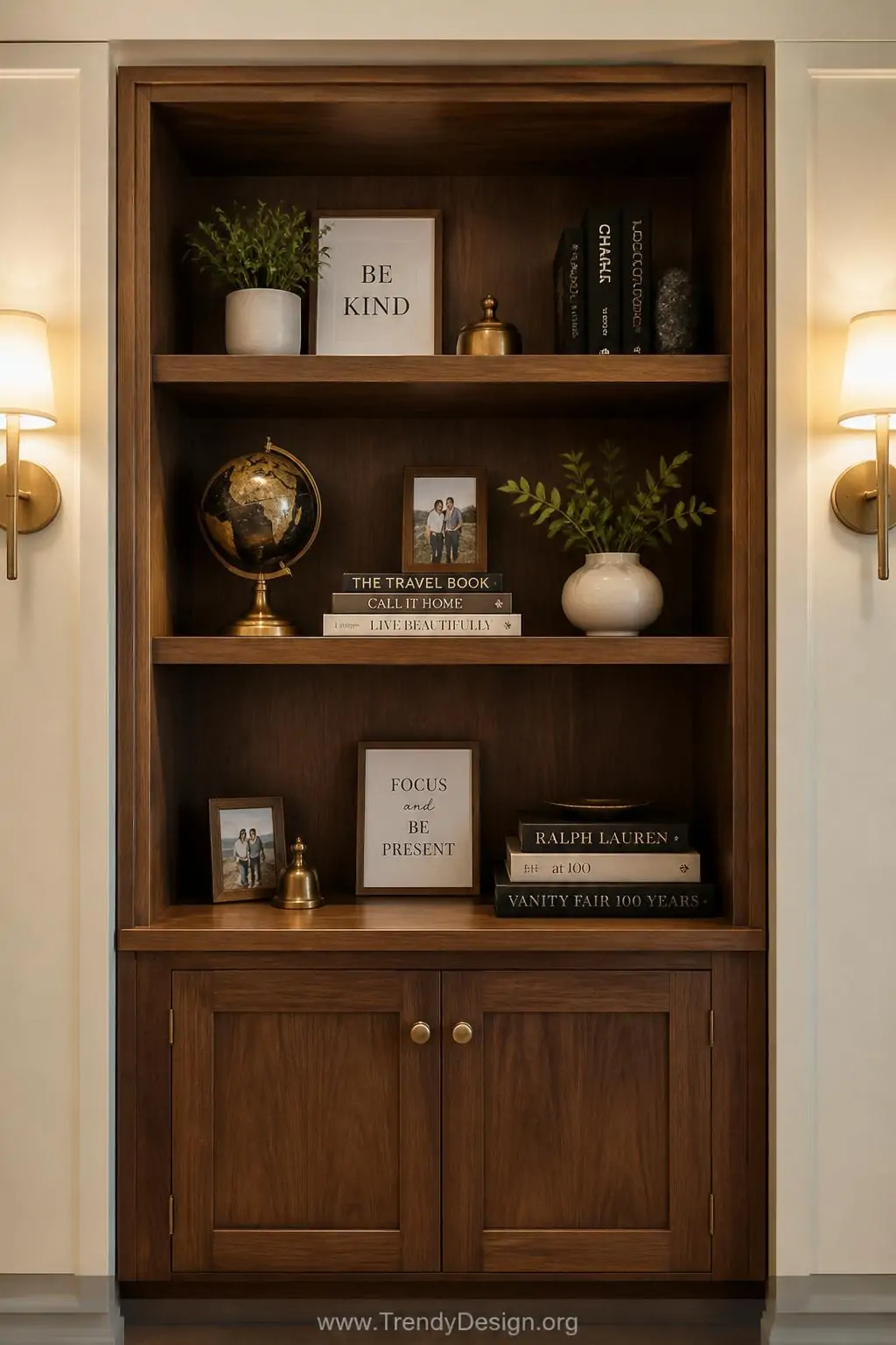 Styled wooden bookshelf vignette displaying a small brass globe, stacked spines of aspirational books, tiny framed motivational prints, and tucked personal photos between curated objects in warm walnut tones against an ivory wall, soft warm sconce lighting illuminating the three-dimensional arrangement, wide shot showing the full shelf composition, photorealistic interior design.